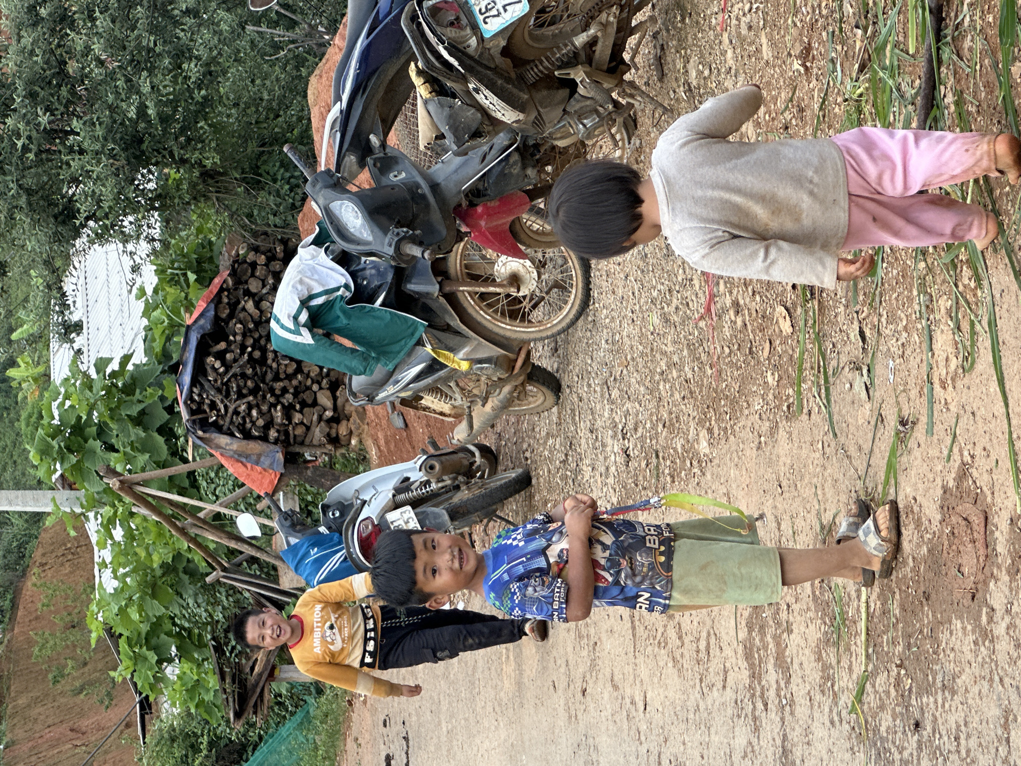This image, taken in Xín Vàng, Vietnam, depicts three children on an unpaved road surrounded by vegetation and village elements.
In the foreground, an unpaved, reddish-brown dirt path with scattered green plant debris extends through the scene. A muddy patch is visible in the lower center.
On the left, an older boy with short dark hair walks towards the right, smiling broadly. He wears a yellow long-sleeved shirt with "AMBITION" and "SPACE SOUL" graphics, dark cargo pants, and dark sandals. Behind him, a light-colored scooter with a partially visible license plate displaying "37 L1 - 53" is parked.
In the center, a younger boy with short dark hair and bangs stands facing the viewer, offering a slight smile. He wears a blue short-sleeved t-shirt featuring multiple Batman characters and light green shorts with checkered pattern slide sandals. He holds a colorful ribbon or strap with a lime green element.
To the right, a third, younger child, seen from behind, walks away from the camera towards the right. This child has short dark hair, wears a light grey long-sleeved top and pink pants, and is barefoot.
In the mid-ground, three motorcycles or scooters are parked. Behind the central boy, a darker motorcycle has a green and white jacket draped over its seat. Further to the right, a heavily dirt-covered dark motorcycle has a partially visible license plate reading "26 T1".
The background features dense green foliage and trees. Through the vegetation, a reddish-brown earthen slope is visible on the far left, and corrugated metal roofs of structures can be seen in the distance. A pile of cut firewood is stacked under a blue and orange tarp on the right, and rudimentary wooden support structures are present in the mid-left. Utility lines are visible in the upper left.