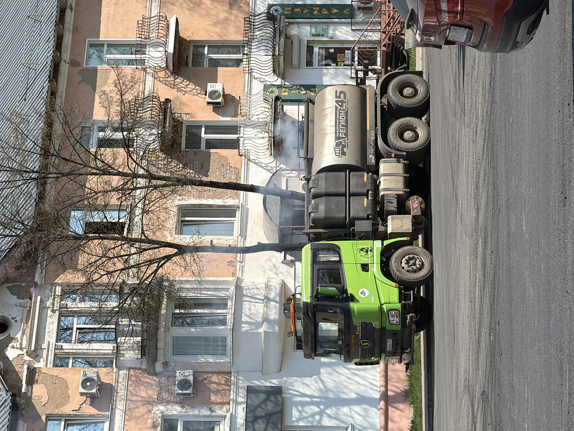 The image shows a green and black tank truck, identified as a "REGION 45" model, positioned on a paved street. The truck is adjacent to a multi-story building with a weathered, textured facade featuring a mix of peach and white sections. Several windows are visible, some with decorative wrought-iron balconies. Air conditioning units are mounted on the building's exterior. A leafless tree branches out in front of the building. Smoke or steam emanates from the truck. In the upper right corner of the image, part of a red vehicle is visible. The location is Votkinsk, Russia, as indicated by the "REGION 45" designation on the truck.