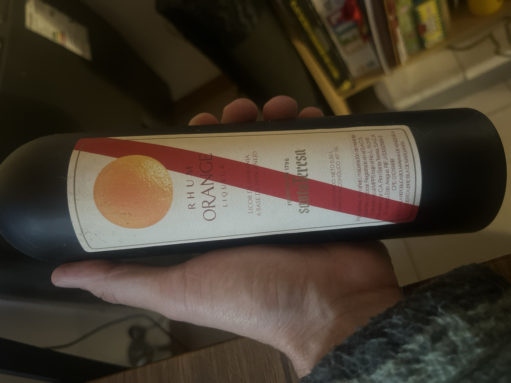 A hand holds a black, cylindrical bottle of "Rhum Orange Liqueur" by Santa Teresa. The label has an orange graphic on the left and a red diagonal stripe across the center. Text on the label reads, in Spanish, "Licor de Naranja a Base de Ron Añejo," and "Contenido Neto 0,50 L Grado Alcoholico 40° GL." There is no indication in the image that it is taken in the city of Ushuaia, Argentina.