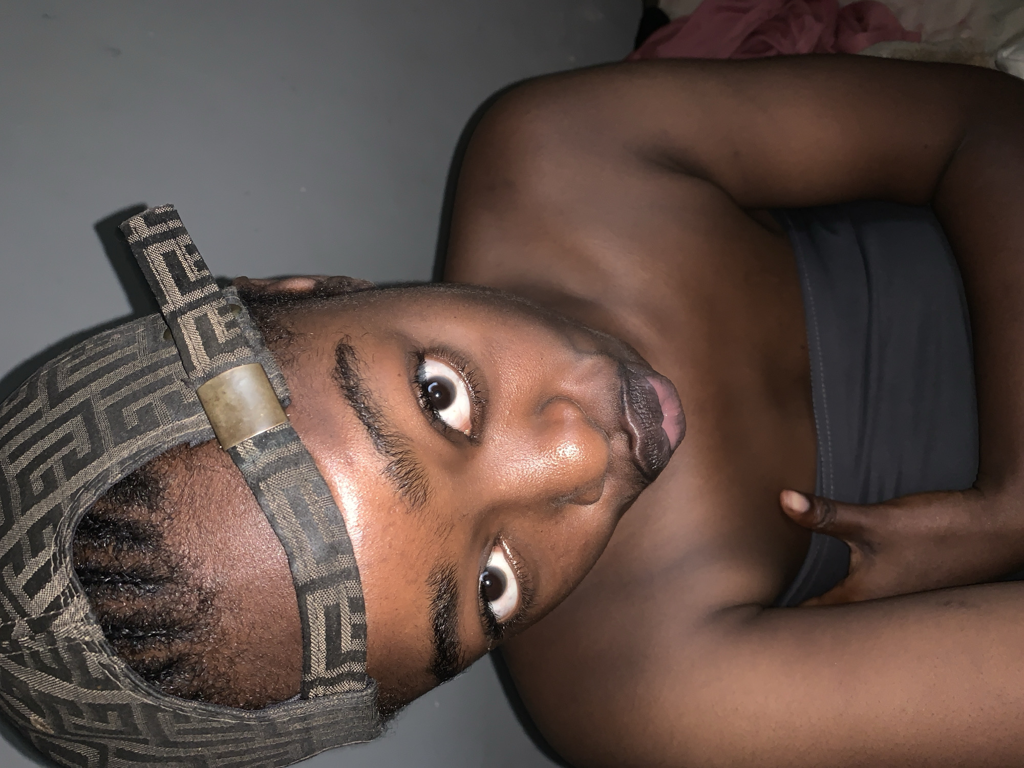 The image shows a close-up, high-angle, point-of-view shot of a young Black person wearing a baseball cap and a strapless top. The person's face is the primary focus, with their eyes looking directly at the camera. The cap has a patterned design. The person's skin appears to have a dewy sheen, suggesting it might be oily or illuminated by light. Their mouth is slightly open, and they are holding their left arm across their chest. The background is a plain, neutral gray wall. The overall lighting is dim, with a slight reddish hue visible on the right side of the image, possibly from clothing or other fabric. No specific location in Rafin Par, Nigeria, is identifiable from the visual elements.
