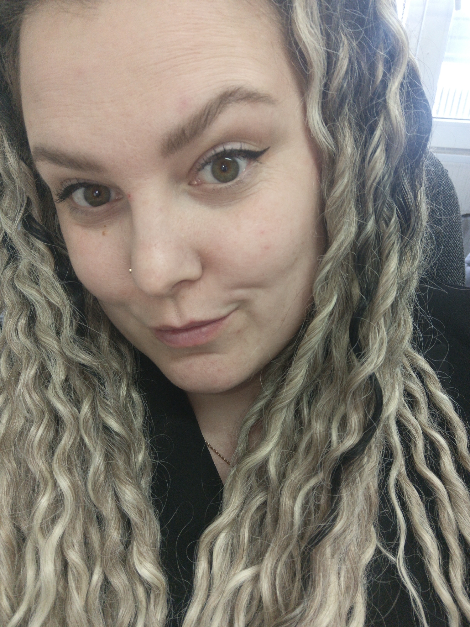 A close-up portrait shows an adult female subject. Her face occupies the majority of the frame, angled slightly toward the viewer. She has long, curly hair with mixed blonde/light brown and darker strands, falling around her face and shoulders. Her eyes are brown-green (hazel), with visible eyeliner on the upper lids. Her eyebrows are dark and full. A small, silver-colored stud piercing is present on her left nostril. Her lips are closed and slightly pursed, indicating a subtle smile. The subject's skin is fair, with fine lines visible on her forehead and a faint mole/freckle below her right eye. She is wearing a dark-colored garment, and a thin, gold-colored chain is visible around her neck. The background is blurred, showing indistinct elements suggesting an indoor environment, including a light-colored rectangular structure, possibly a window, and a darker, textured surface. The subject is looking directly at the camera. The specific city of Noviy, Russia, cannot be determined from the visual information within the image.