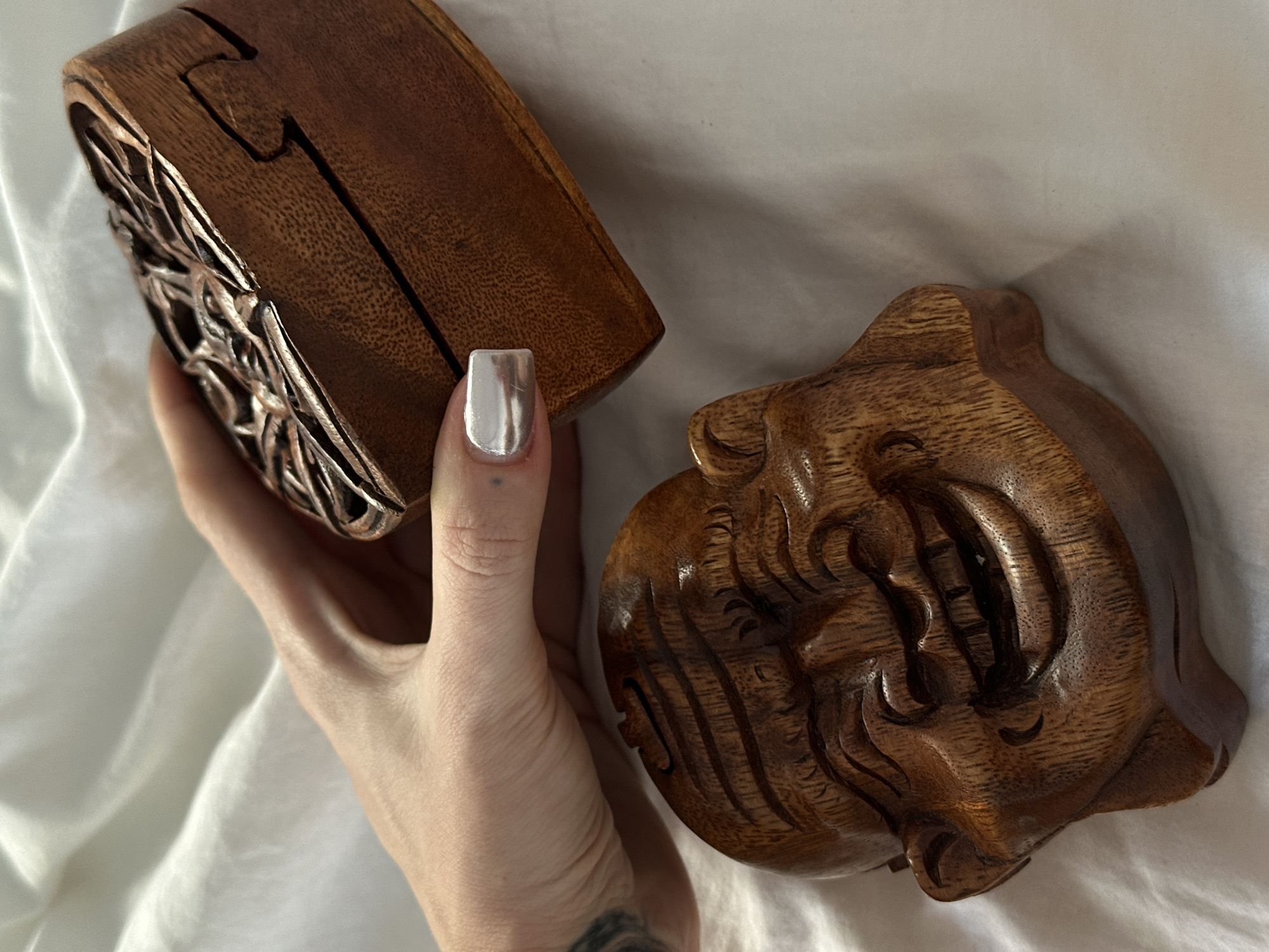 A person's hand, adorned with metallic silver nail polish, interacts with two intricately carved wooden objects, set against a backdrop of soft white fabric. The hand holds a dark brown wooden box, possibly a puzzle box, which features elaborate, openwork carvings resembling foliage or abstract patterns on its side. Visible seams on the box suggest a hidden opening or mechanism. Next to the hand, lying on the white fabric, is another piece of carved wood, also dark brown, shaped like a stylized, almost mask-like face or head with deep, swirling grooves and defined features, hinting at a mythical creature or traditional design. A tattoo is partially visible on the person's forearm near the wrist. The lighting is even and soft, highlighting the natural wood grain and the detailed craftsmanship of both pieces. Given the context of North Kuta, Indonesia, these items are likely examples of traditional Balinese wood carving.