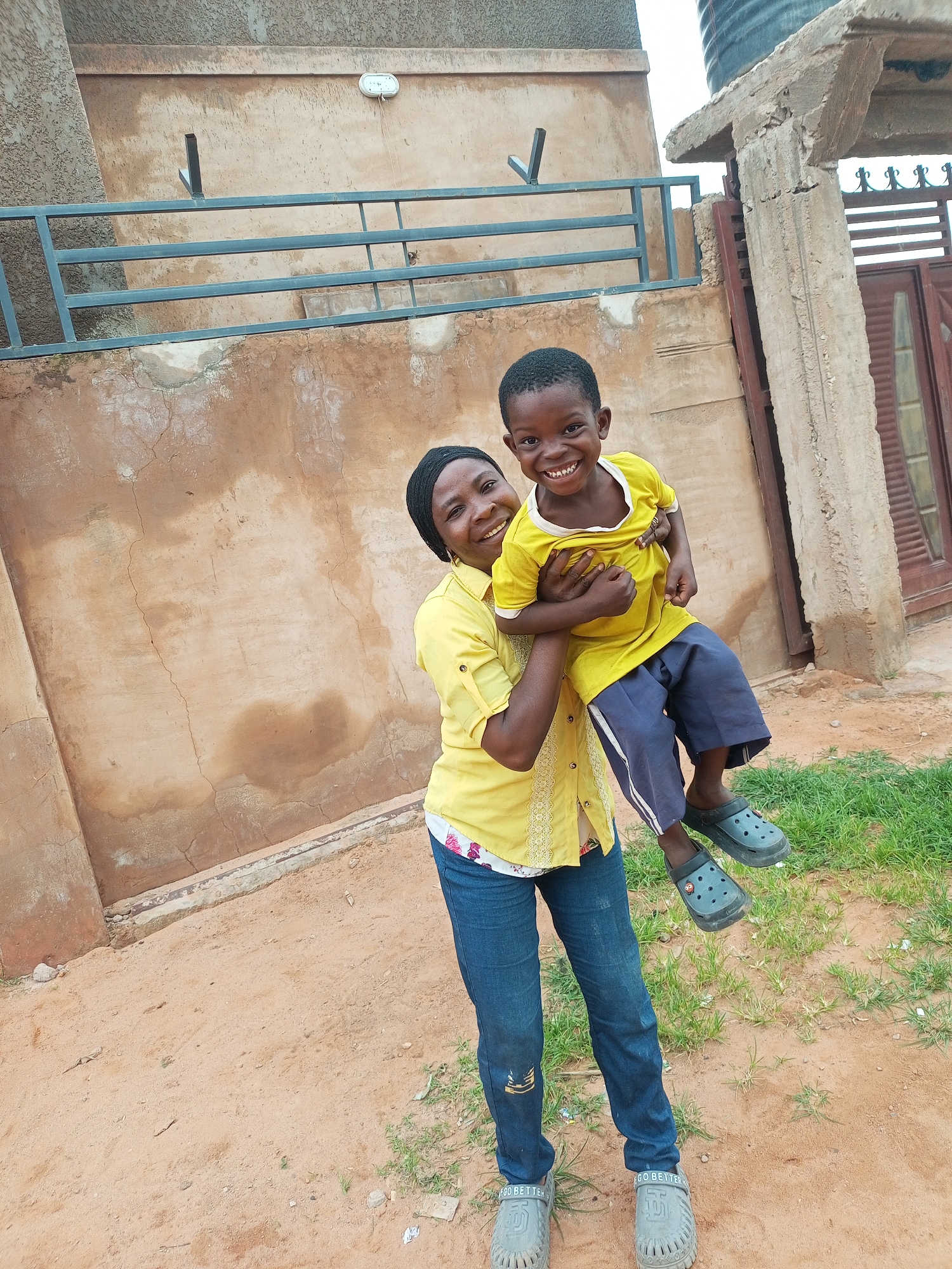 A smiling woman wearing a yellow shirt and blue jeans holds a smiling young boy in her arms. The boy is wearing a yellow t-shirt and blue shorts with white stripes. Both are wearing gray clogs. They are standing in front of a textured, light brown wall. To the left of the wall is a dark metal railing. To the right of the wall is a concrete structure with a dark brown door and a large black water tank on top. The ground is covered in sand and patches of green grass. The scene appears to be outdoors in Tempure, Nigeria.