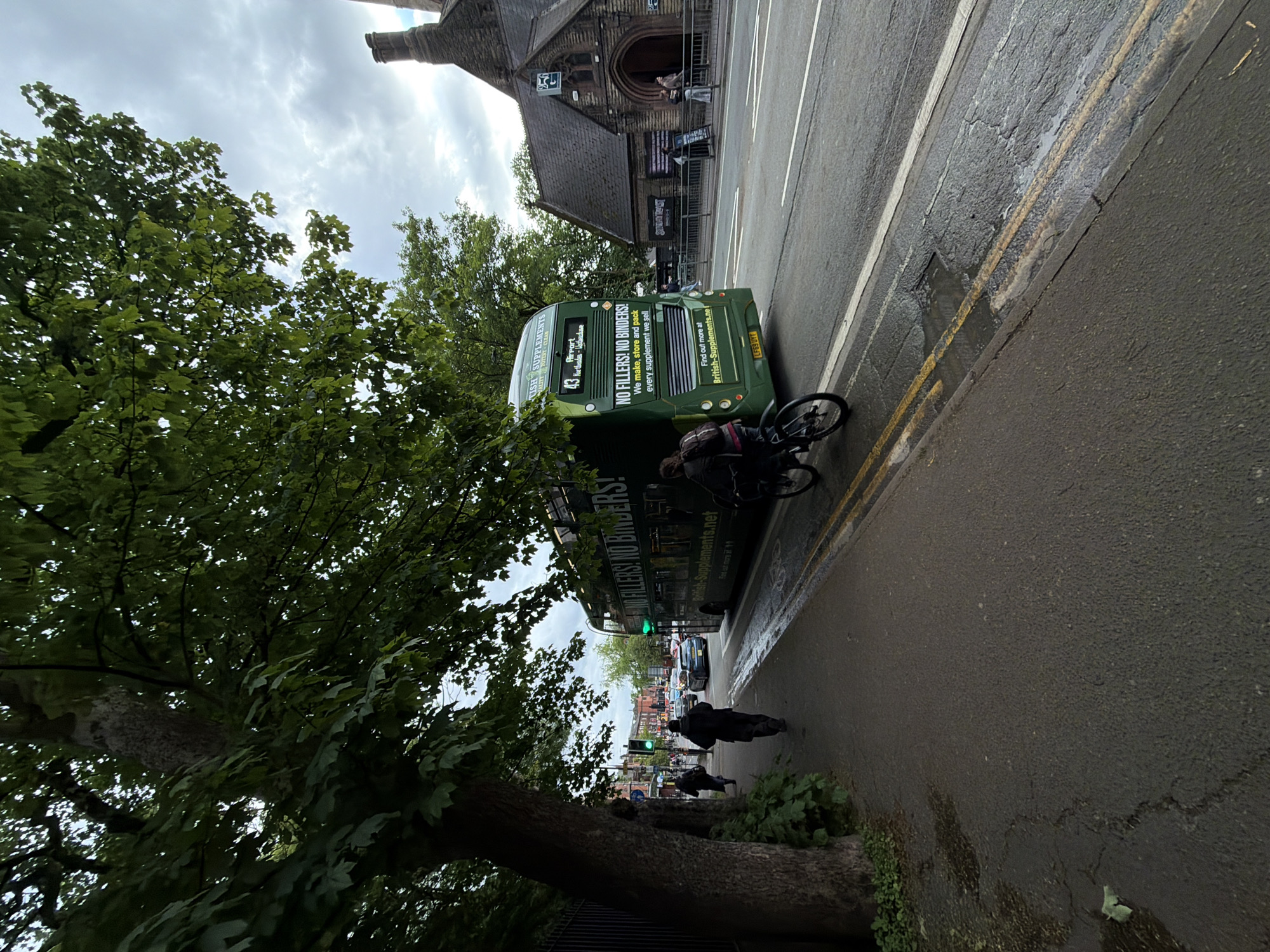 The image depicts a street scene in an urban environment. A large green double-decker bus with the advertisement "NO FILLERS! NO BINDERS!" occupies a significant portion of the frame, parked near the curb. A cyclist is near the bus. The street has a yellow line. On the left is a large tree, partially obscuring a building. Pedestrians are visible on the sidewalk. Traffic lights are seen down the street. The sky is cloudy. The building in the background has a distinctive architectural style, with a tower-like structure.