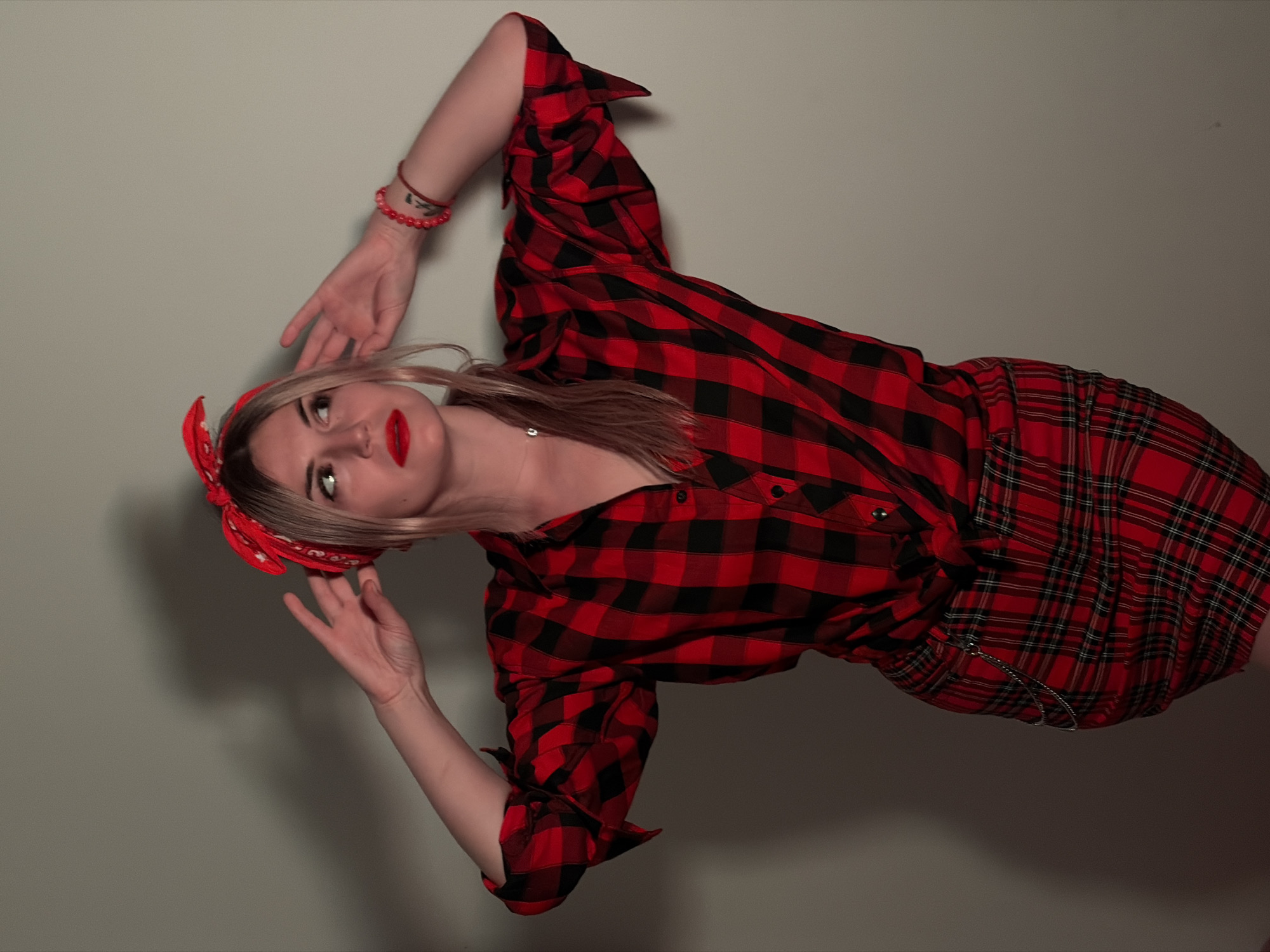 A woman is posing indoors against a light grey wall. She is wearing a red and black plaid shirt and skirt, with a matching red headband. She has blonde hair, red lipstick, and is wearing a red beaded bracelet on her left wrist. Her arms are raised, with her hands slightly open. A thin silver chain is attached to her skirt. The image is rotated slightly counter-clockwise. Budva, Montenegro cannot be determined from the provided image.
