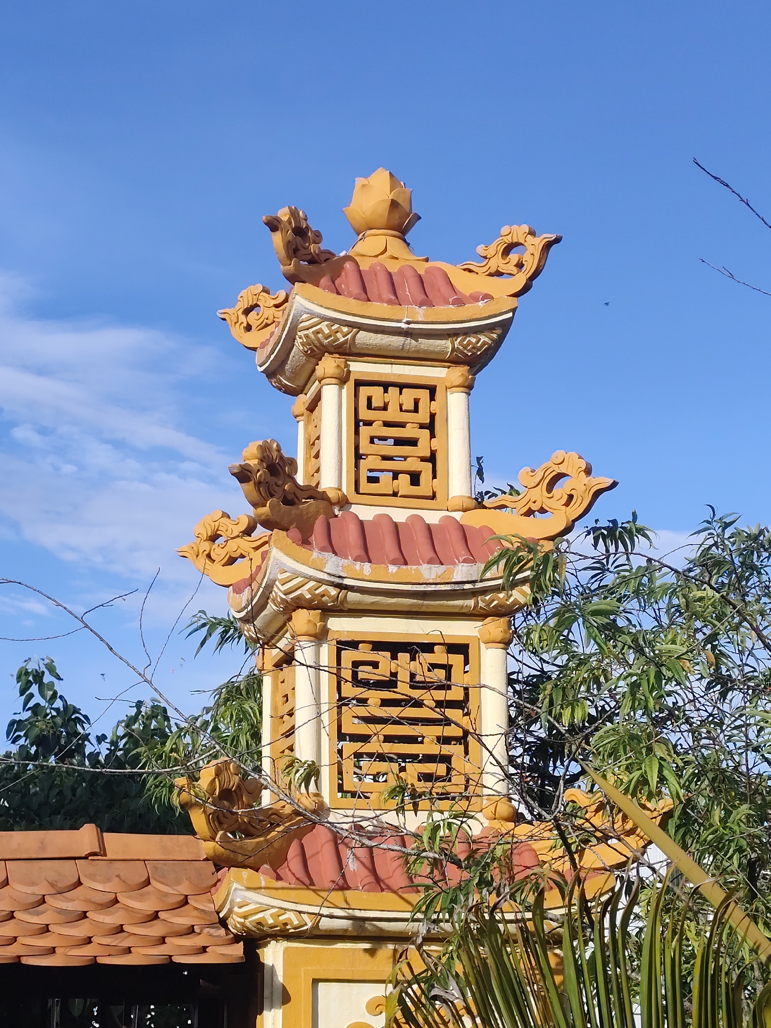 This image captures a close-up view of the ornate upper section of a multi-tiered pagoda, likely located in Da Lat, Vietnam. The structure is predominantly yellow and red, with intricate decorative elements. Each tier features a red, curved roofline with prominent yellow, sculpted embellishments that resemble dragon heads or mythical creatures. The central body of each tier is a square pillar with openwork panels, revealing a complex geometric pattern in a dark color against a yellow background. At the very top, a golden lotus bud finial crowns the structure.
The pagoda is set against a clear blue sky with a few wispy white clouds. Green foliage from trees partially obscures the lower portions of the pagoda, suggesting it's situated within a natural or garden setting. The presence of bare branches reaching into the frame adds a touch of natural detail. The lighting indicates it's daytime, likely morning or afternoon, with bright sunlight casting shadows and highlighting the textures of the building and the leaves. There is no visible text in the image. The overall impression is one of architectural beauty and cultural significance, set within a peaceful, outdoor environment.