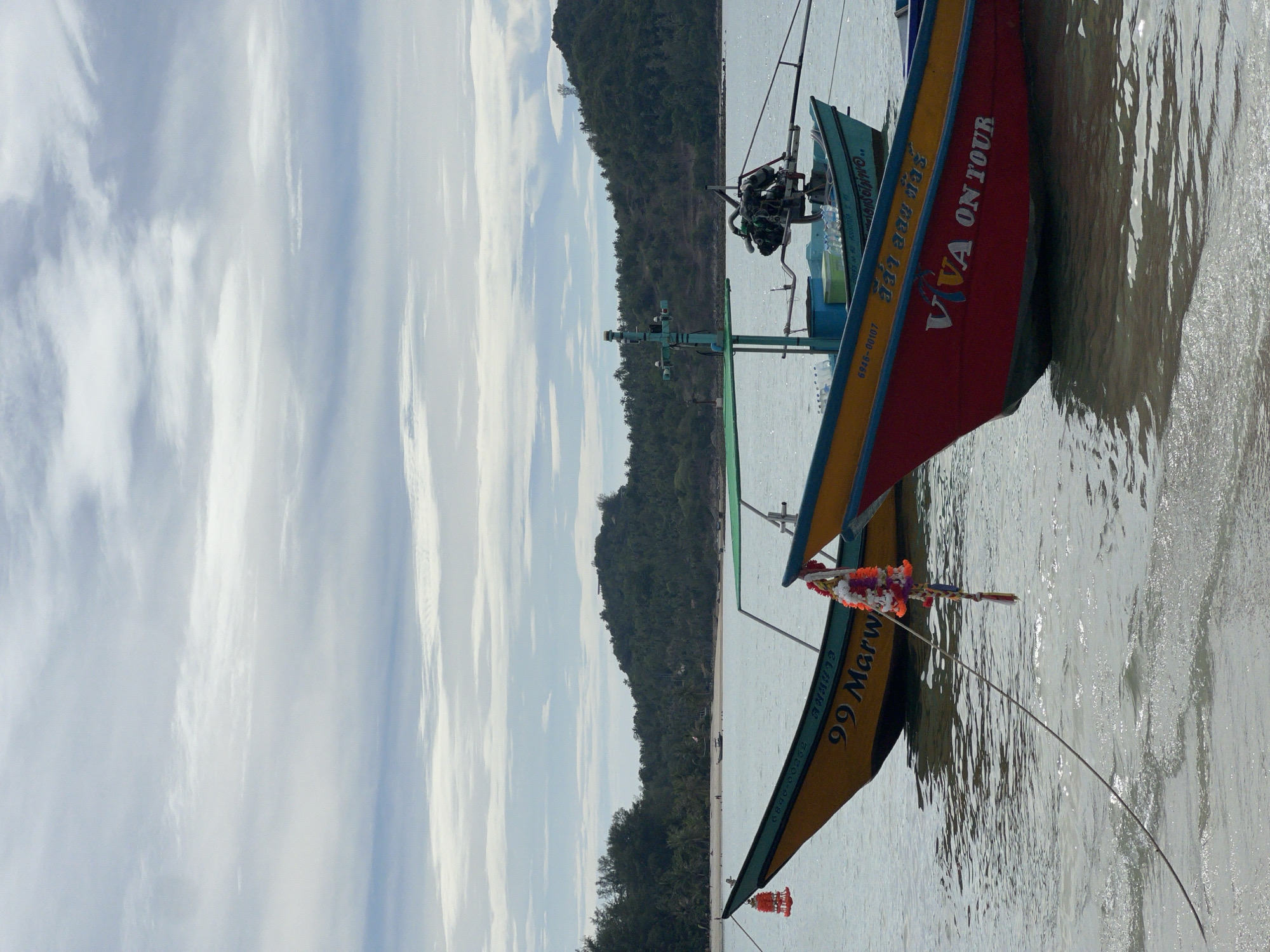 The image depicts a serene scene on the water in Koh Phangan, Thailand. Two colorful longtail boats are anchored close to the shore, their hulls painted in vibrant hues of yellow, red, and blue. The boat in the foreground, with its striking red hull, bears the inscription "VIVA ON TOUR" in white lettering, suggesting it's a vessel used for tourist excursions. The yellow boat to its left displays the number "99 Marw" and a registration code. Both boats are adorned with decorative garlands. The water is calm and shallow near the shore, with gentle ripples reflecting the overcast sky. In the background, a lush, green, tree-covered hill slopes down to a sandy beach dotted with a few distant figures. The sky is a mix of white and gray clouds, suggesting a possibly humid or rainy day. The overall atmosphere is peaceful and tropical.