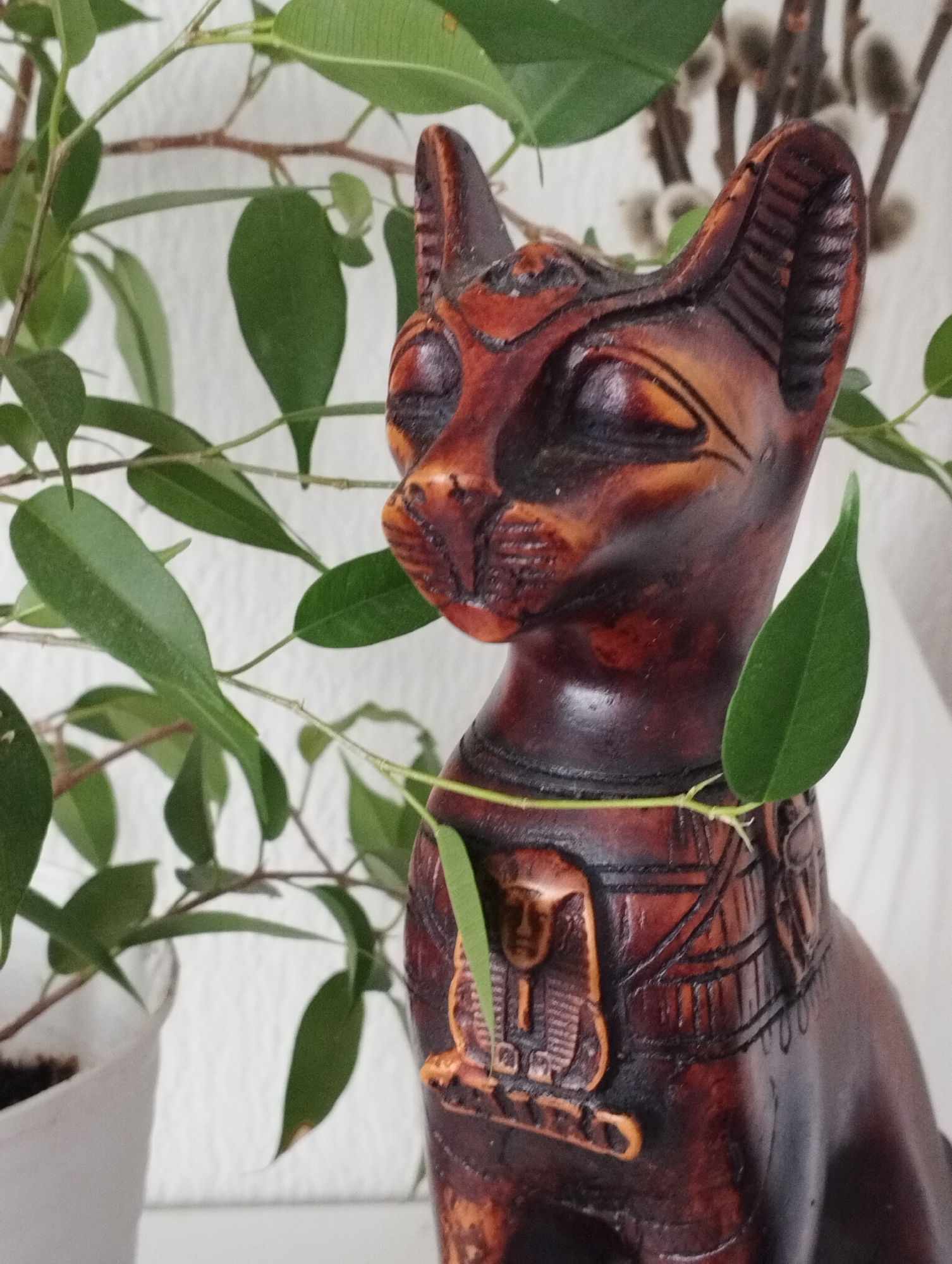 A carved, dark brown Egyptian Bastet cat statue stands amidst the foliage of a potted plant. The statue, likely made of wood, features intricate carvings depicting an Egyptian pharaoh on its chest. A potted plant sits to the left, its green leaves partially obscuring the statue. Behind the statue and plant is a white wall. In the background, slightly out of focus, are some pussy willow branches. No people are visible. There is no information available about the city Sadovyy, Russia, or its relation to this image.