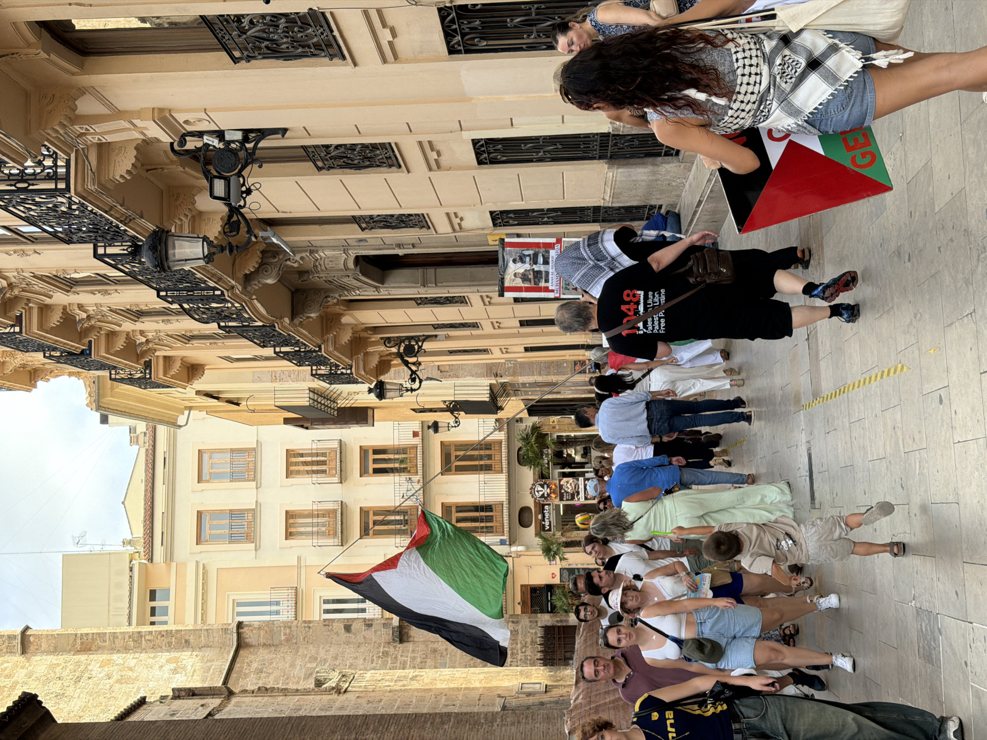 In an urban environment, a group of people are walking down a paved street lined with buildings. A large Palestinian flag is being held aloft on a pole, diagonally across the scene. Several individuals are visible, some walking in the same direction, others in different directions. One person in the foreground, on the right, is carrying a triangular flag featuring the Palestinian flag's colors and the word "GE...". Another person, on the left, is wearing a black t-shirt with white text that includes "1948". A young child in a beige t-shirt and shorts is walking away from the camera. The buildings flanking the street appear to be old, with balconies and architectural details. The sky is overcast.
