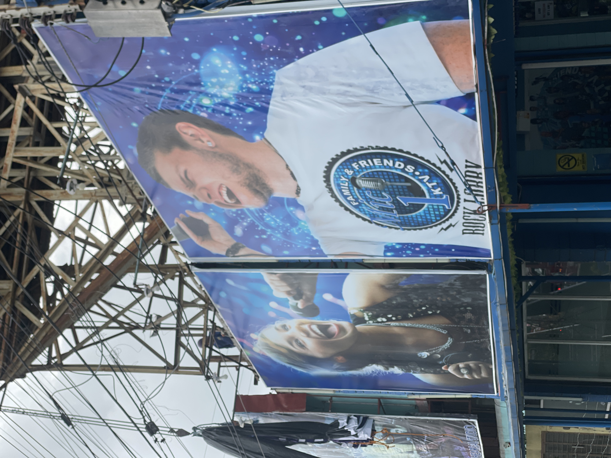 A medium shot captures a billboard in a city, likely Davao City, Philippines, promoting a KTV establishment. The billboard features two prominent images: on the left, a woman with her mouth wide open, holding a microphone and wearing a sparkly top; on the right, a man with his eyes closed and mouth open, also holding a microphone and wearing a white t-shirt with a circular logo that reads "FAMILY & FRIENDS Mic 1 KTV". Below the images, the text "ROCK LARRY" is visible. The billboard is positioned above a building with a blue facade and glass windows. Overhead, a complex network of electrical wires and metal scaffolding obscures the sky, suggesting an urban environment. The lighting indicates it is daytime, with an overcast sky.