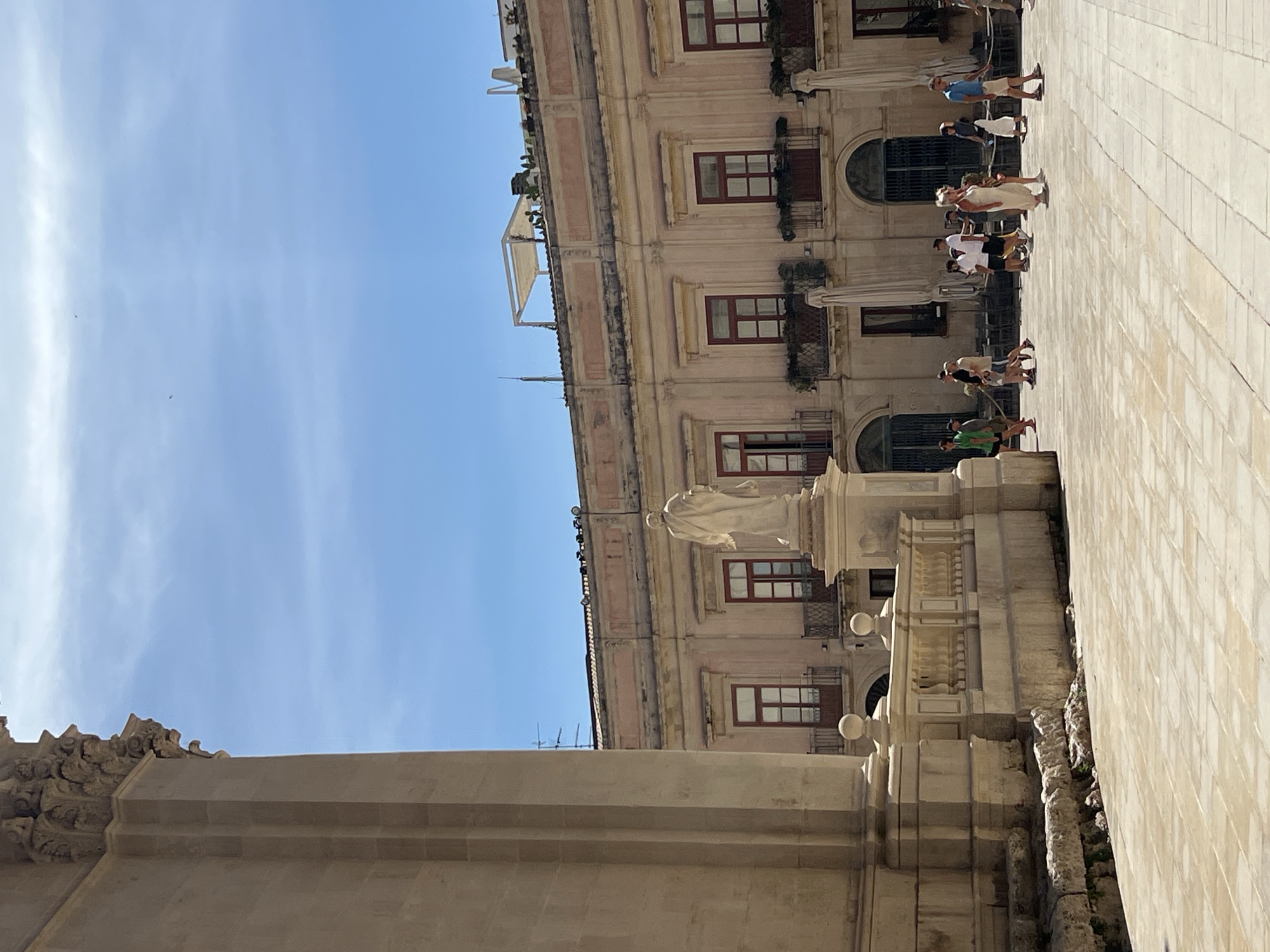 The image depicts a sunlit piazza in Syracuse, Italy, characterized by its historic architecture and scattered groups of people. In the foreground, a large, light-colored stone column dominates the left side of the frame, suggesting the presence of a significant building, likely a cathedral or ancient structure, given the elaborate carvings at its top. The ground is paved with large, light-colored rectangular tiles, indicative of a public square.
Dominating the midground is a pale pink building with multiple stories, featuring red-framed windows and decorative balconies. A prominent white statue of a robed figure stands on a pedestal in front of this building, facing away from the viewer. Several individuals are captured in motion across the piazza. To the right of the statue, a diverse group of tourists strolls casually. They are dressed in light summer clothing, including dresses, shorts, and t-shirts. Some are walking individually, while others are in small clusters. The overall atmosphere suggests a typical day in a tourist-friendly historical location, with clear blue skies and bright sunlight casting shadows. The presence of umbrellas, possibly from outdoor cafes, and the architectural style strongly suggest a Mediterranean setting, consistent with Syracuse.