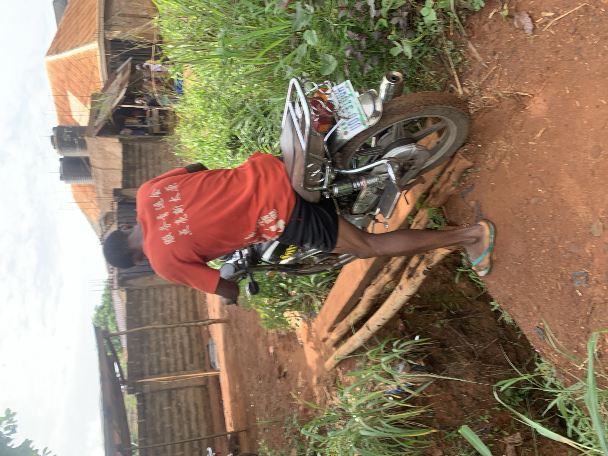 The image shows a man in Ukwu-Abwa, Nigeria, standing next to a motorcycle that has fallen onto its side. The man is wearing an orange t-shirt and black shorts with flip-flops. The motorcycle's rear wheel is visible, along with a license plate. The environment includes a dirt path, green vegetation, and a building in the background.