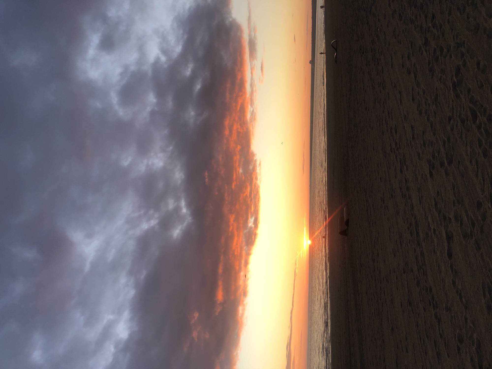 This photograph captures a sunset over the Gulf of Finland in Sestroretsk, Russia. The sky is dominated by dramatic clouds, with the lower portions illuminated by the setting sun in fiery oranges and reds, transitioning to darker grays and purples higher up. The sun itself is a bright, low-hanging disc on the horizon, casting a strong glare across the water. The sea is calm with subtle ripples. The foreground is a sandy beach, appearing dark due to the low light, with visible footprints. Scattered across the beach are a few individuals: a person lying down in the middle ground on the left, and several distant figures walking or standing near the waterline on the right. A dark, curved object, possibly a piece of driftwood or debris, lies on the sand in the right foreground. In the distance, faint silhouettes of buildings are visible along the horizon.
