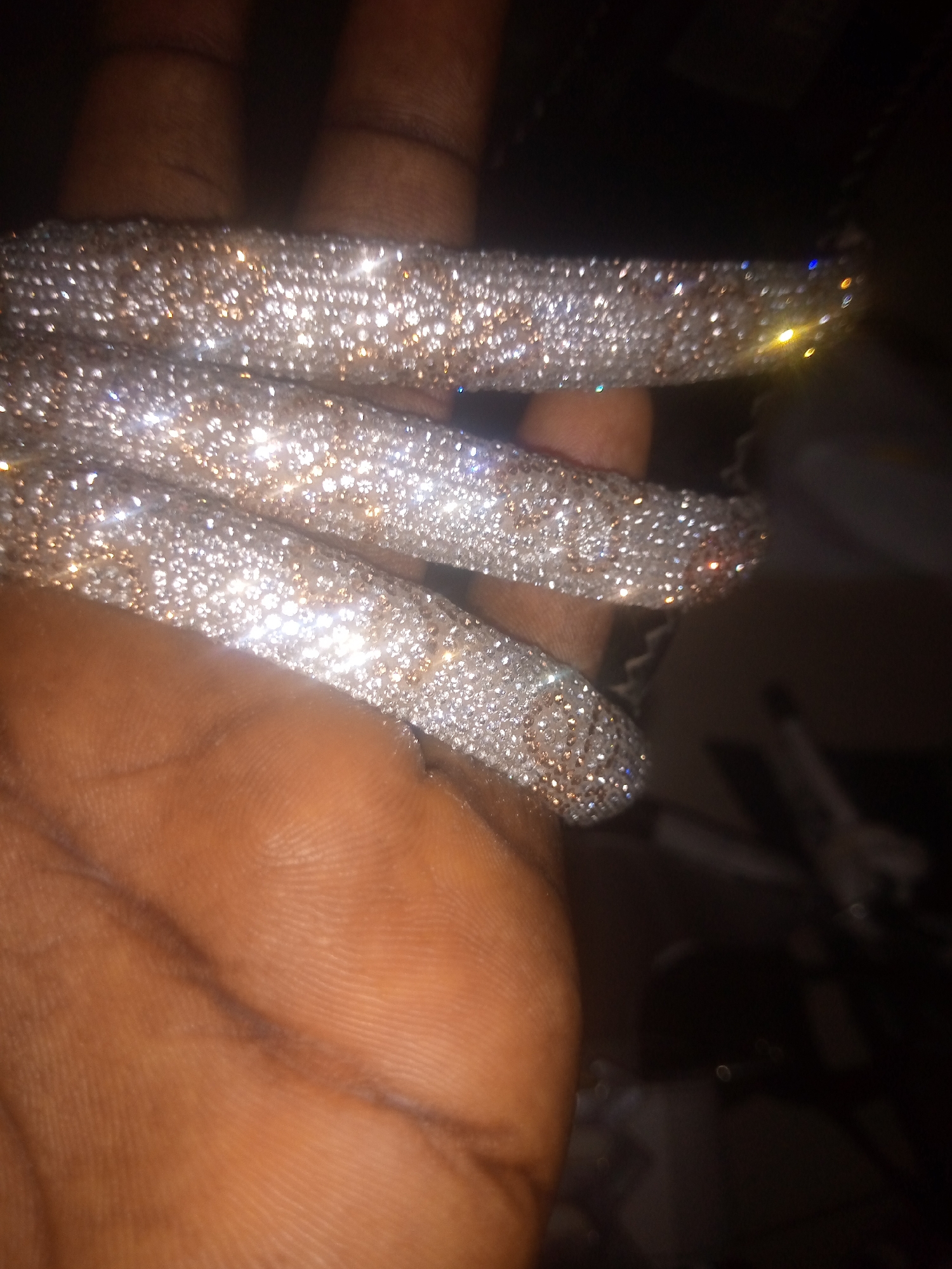 The image is a close-up, overhead shot of a hand adorned with long, bejeweled artificial fingernails. The nails are covered in a dense pattern of small, shimmering rhinestones, giving them a sparkling, diamond-like appearance. The hand itself is dark-skinned, and its texture, including fingerprints, is visible. The background is dark and out of focus, suggesting a dimly lit environment, possibly indoors. There are some faint, out-of-focus lights in the background, some of which are colored, indicating artificial lighting sources. The image does not provide any specific information about the location in the city of Jauro Yinu, Nigeria.