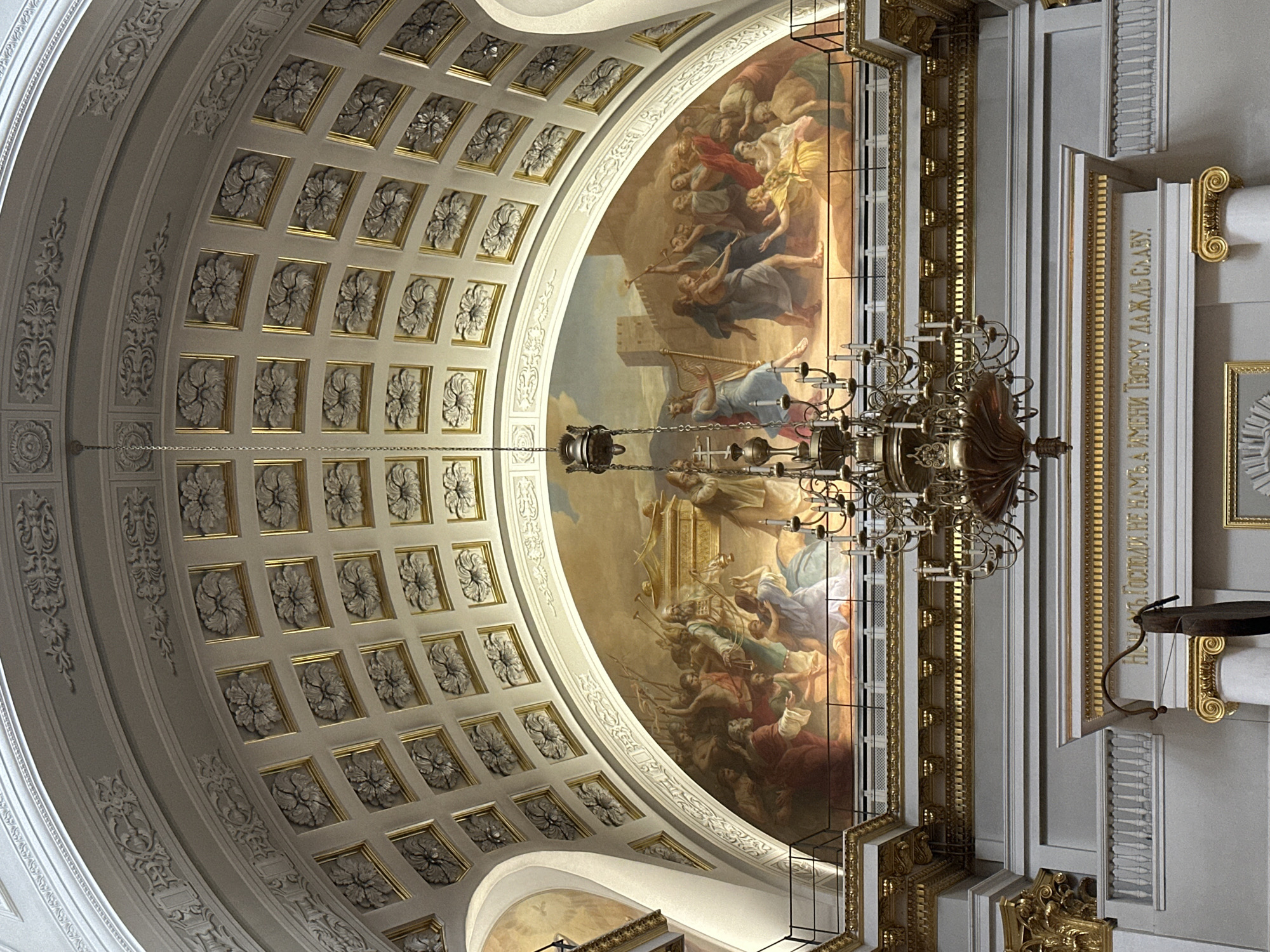 The image shows the interior of a cathedral, likely in Kazan, Russia, focusing on the ornate ceiling. The ceiling is divided into two main sections: one side is decorated with repeating square panels featuring floral motifs in a grid pattern, each panel edged in gold. The other side is covered by a large, curved fresco depicting a historical or religious scene with many figures, some in robes and others in what appears to be battle.
A large, multi-tiered chandelier hangs in the center of the frame, partially obscuring the fresco. The architectural details are emphasized by gold trim and moldings. A portion of a wall with inscriptions in Russian is visible to the right, indicating it's a religious or historical site. The overall impression is that of a grand and opulent space, reflecting a significant cultural or religious heritage.