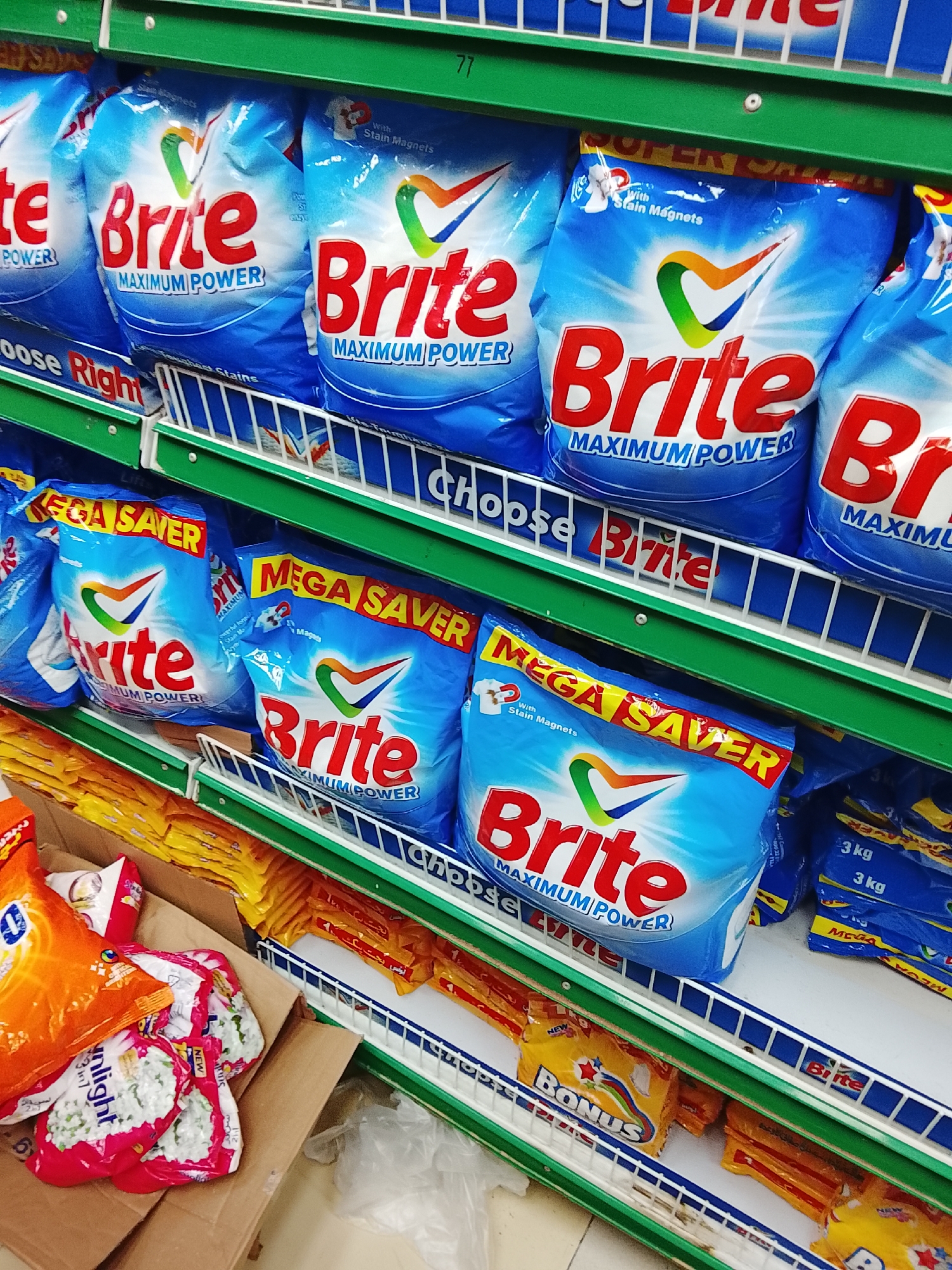 The image shows a shelf in a store, likely a grocery or convenience store, in Gulgasht Colony, Pakistan. The shelves are green and white, stocked with various laundry detergent products. On the top shelves are multiple blue bags of "Brite Maximum Power" laundry detergent. Below those are smaller bags of "Brite Mega Saver" laundry detergent. The bottom shelf contains yellow bags of "Bonus" dishwashing liquid and orange and red bags of "Sunlight" detergent. The perspective is from a close vantage point, focusing on the detergents.