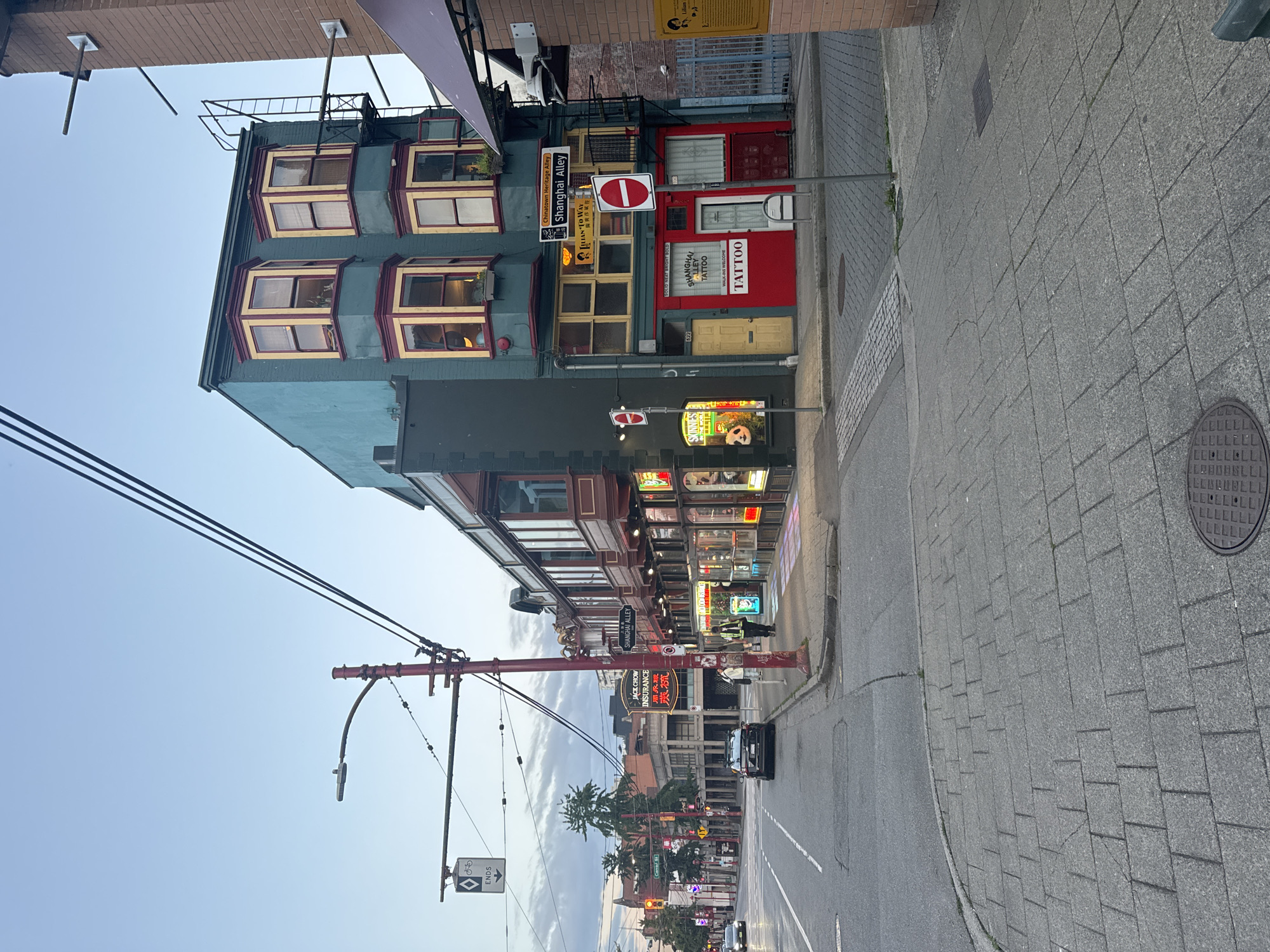 The image captures a street scene in Vancouver's Chinatown district, specifically on Shanghai Alley. The time appears to be late afternoon or early evening, with a soft, diffused light. The architecture is a blend of older, heritage buildings with distinct bay windows and more modern elements. On the right, a teal-colored building stands prominently, featuring several yellow-trimmed bay windows on its upper floors and a bright red facade on the ground level. Signs indicate a tattoo parlor, "Solid Neat Bright Bold," and a business named "Lilian-to-Way." Further down the street, neon signs glow, advertising businesses such as "Jack Chow Insurance."
The street itself is paved with asphalt, with a section of textured paving leading up to a sidewalk. The sidewalk is made of grey interlocking bricks. Several streetlights and utility poles are visible, some adorned with red decorative elements. A red-painted lamppost with intricate detailing stands on the corner. Traffic signs, including a "No Entry" sign and a sign indicating "Chinatown Heritage Alley Shanghai Alley," are present.
A few individuals are on the sidewalk. One person, wearing a high-visibility vest, appears to be a city worker or security guard. Another person, in lighter clothing, is walking. A black sedan is parked along the curb on the left side of the street, and another car is visible in the distance. The overall atmosphere is that of a bustling urban environment, likely a commercial area with residential units above. The sky is a pale blue with some clouds, suggesting a transition from day to evening.