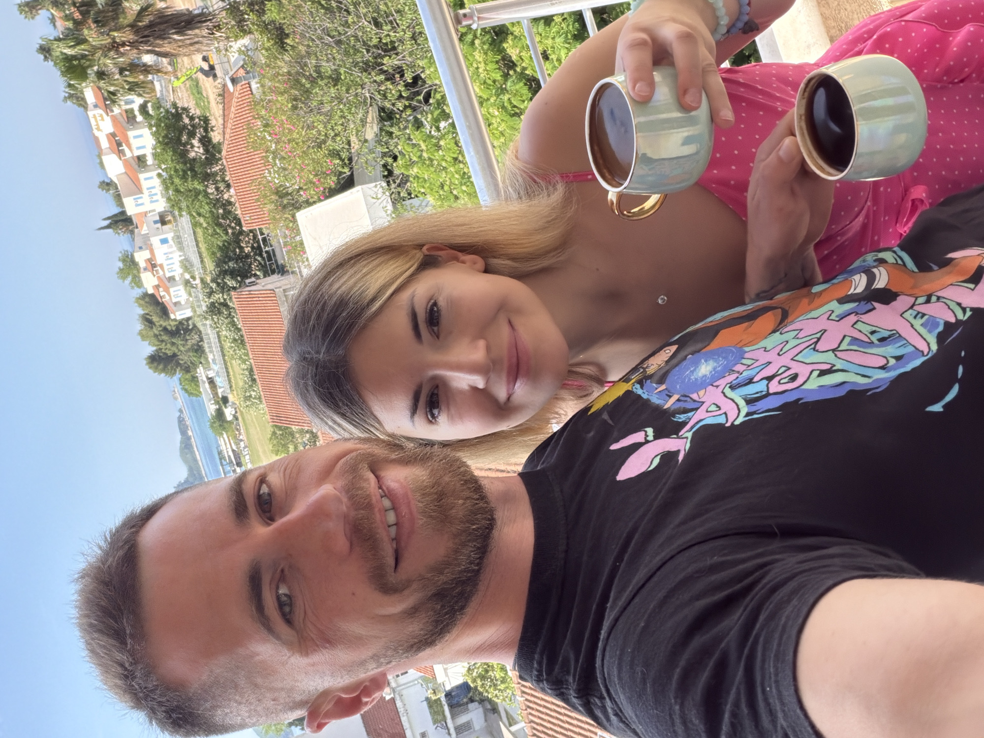 A selfie-style photograph captures a man and a woman posing with two coffee cups. The man, positioned in the foreground, has a beard and is wearing a black t-shirt with a multicolored graphic. The woman, behind him, has blonde hair and is wearing a pink top with white polka dots. Both individuals are holding light blue mugs with gold handles, seemingly containing coffee.
The background reveals a daytime cityscape featuring buildings with red tiled roofs, greenery, and a glimpse of the sea. The image was taken on a balcony, with a railing visible. The location is Budva, Montenegro.