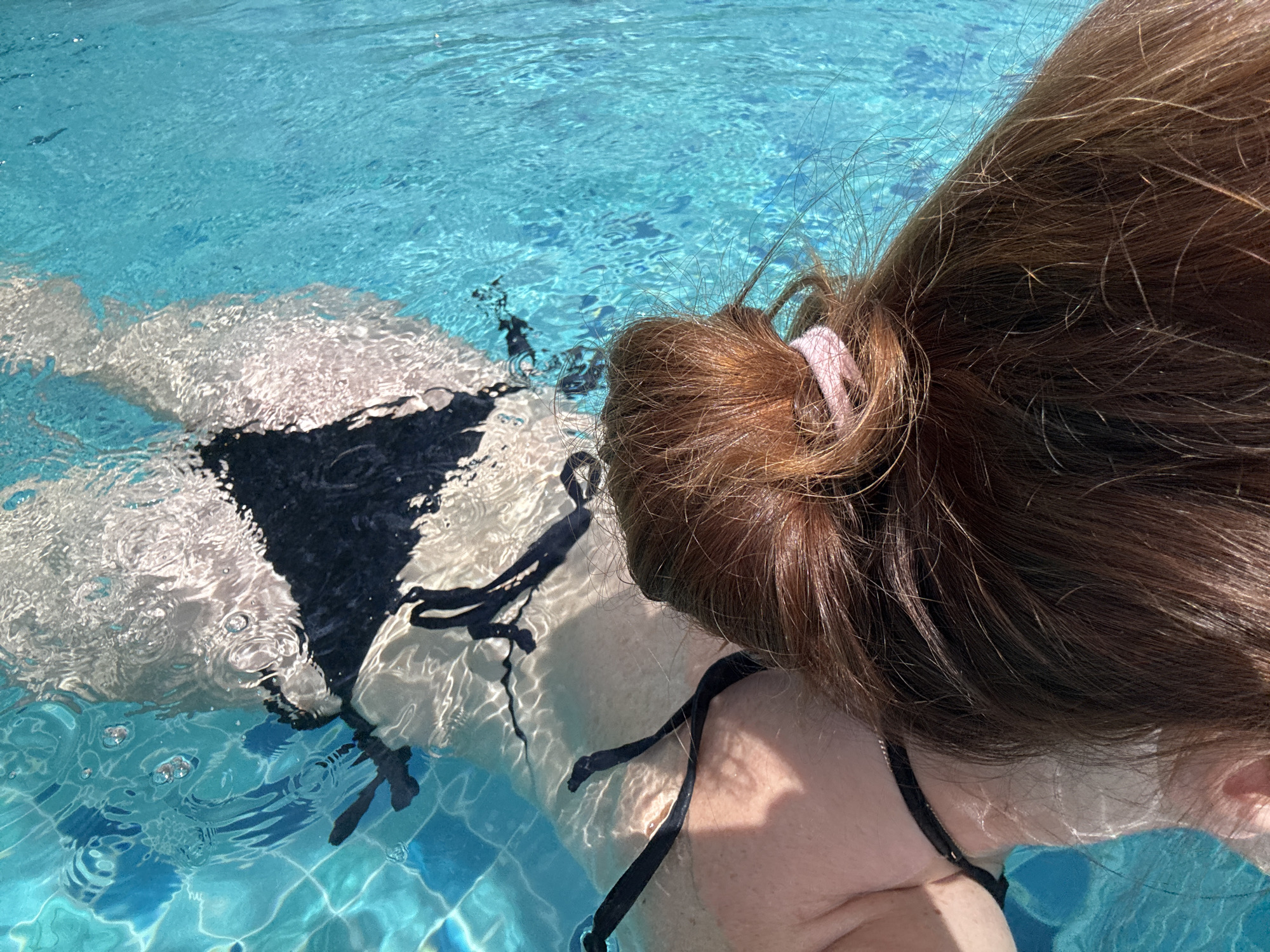A close-up, over-the-shoulder view shows a person in a swimming pool in Baan Plai Laem, Thailand. The person is partially submerged in the turquoise water, with their back to the camera. They are wearing a black bikini. Ripples and light reflections distort the view of their body and the tiled bottom of the pool. The person's long, reddish-brown hair, tied back with a pink scrunchie, dominates the foreground.