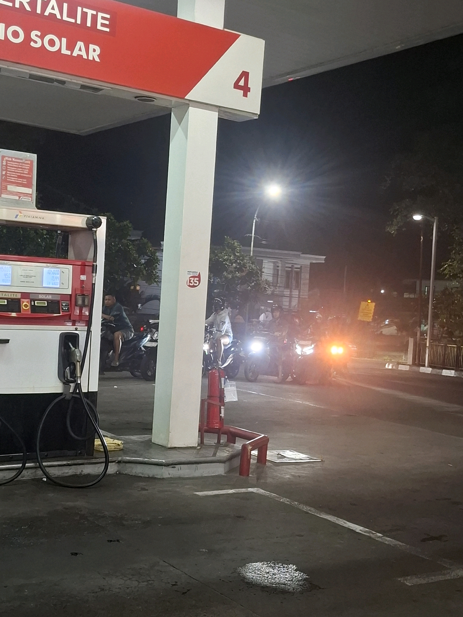 The image is a nighttime, medium-angle shot of a Pertamina gas station in Karangtengah, Indonesia. A white and red gas pump is prominently featured on the left, with digital displays showing fuel prices and quantities. A black hose and nozzle are visible, attached to the pump. Several people on motorcycles are present, some with their headlights on, creating bright flares in the image. One person is seated on a motorcycle next to the pump, while others are further back on the road. Buildings and trees are visible in the background. Streetlights illuminate the scene, casting light and shadows. The ground appears to be wet in places. A red fire extinguisher is mounted on a white pole to the right of the pump. A red and white sign with the number "4" is attached to the overhanging structure of the gas station. Another red circular sign with the number "35" is visible on a white pillar.