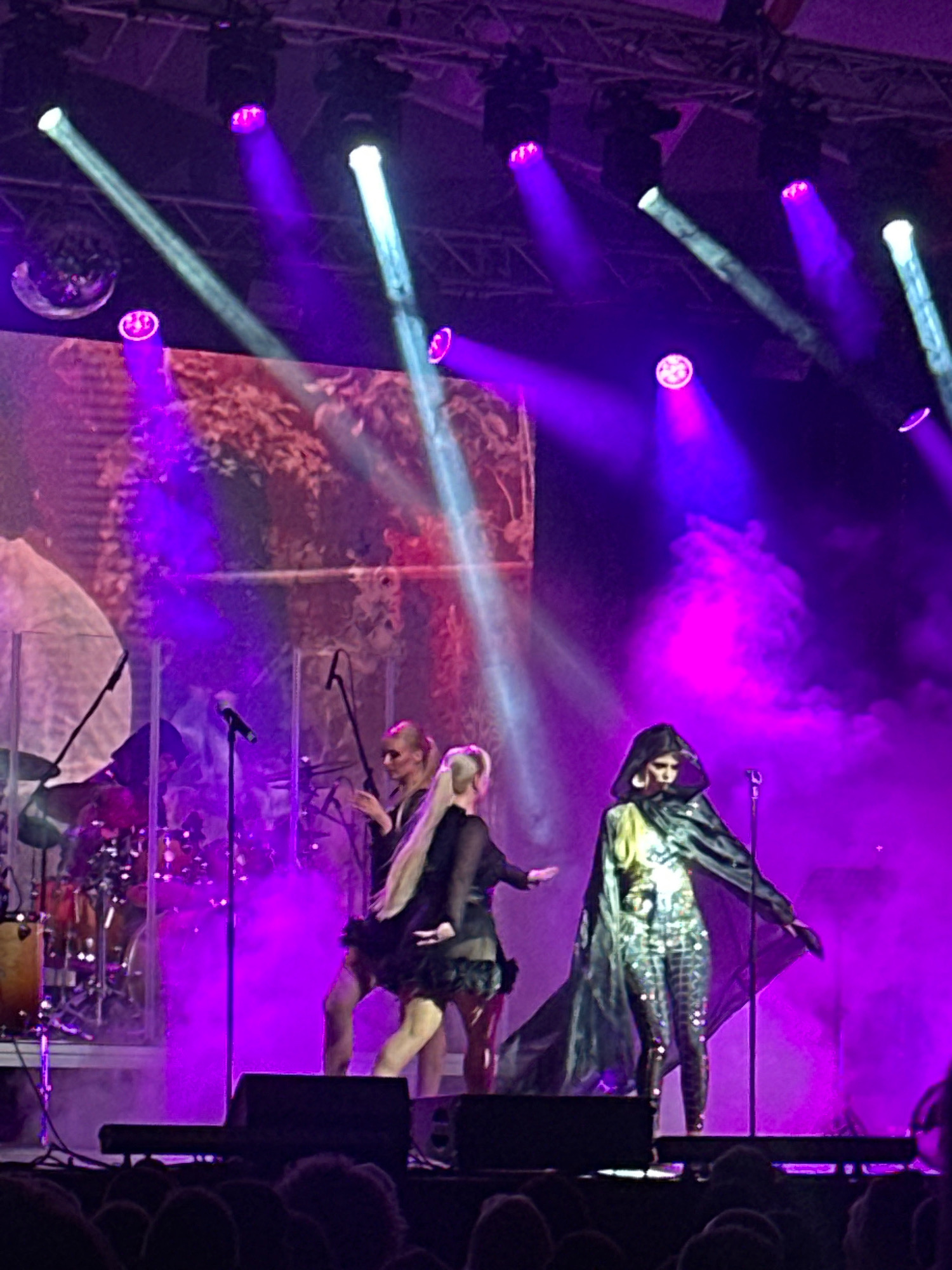 The image depicts a concert or performance on a stage illuminated by purple and white lights. In the foreground, several performers are visible. On the right, a person in a dark hooded cloak and sparkly leggings is standing. To their left, another performer with long blonde hair is seen from the back, wearing a black dress. Further to the left, another individual with blonde hair pulled back in a ponytail is standing, facing away from the camera. Behind the performers, a drum set is partially visible, along with microphones and stage equipment. The backdrop appears to be a large screen displaying an image, possibly of foliage. The overall atmosphere is theatrical and vibrant, with smoke or haze contributing to the lighting effects. The image is taken from a lower angle, suggesting the viewer is part of the audience. The specific location in Jūrmala, Latvia, is not discernible from the visual information provided.