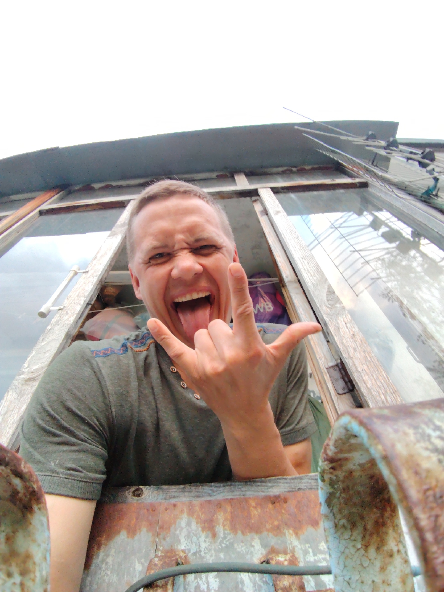 This low-angle portrait, taken in Chaikovsky, Russia, features a man captured in a moment of playful exuberance. He has short hair and is wearing a dark green short-sleeved polo shirt with a decorative, patterned trim near the collar. His mouth is open in a wide grin, with his tongue sticking out, and he is making the "rock on" hand gesture (corna) with his right hand, conveying a sense of energy and fun.
The man appears to be looking out from an open window or aperture of a rustic building. The immediate surroundings consist of weathered wooden frames and glass panels, reflecting an outdoor scene that might include a structure like a greenhouse or another building. In the upper right background, against a bright, overcast sky, parts of an antenna or similar metallic fixture are visible. Behind the man's left shoulder, a purple bag with the letters "WB" clearly visible on it can be seen through the window. In the foreground, heavily rusted and peeling metal surfaces, possibly part of a railing or the structure's exterior, dominate the bottom of the frame, hinting at an aged or industrial setting. The bright, diffuse lighting suggests it is daytime.