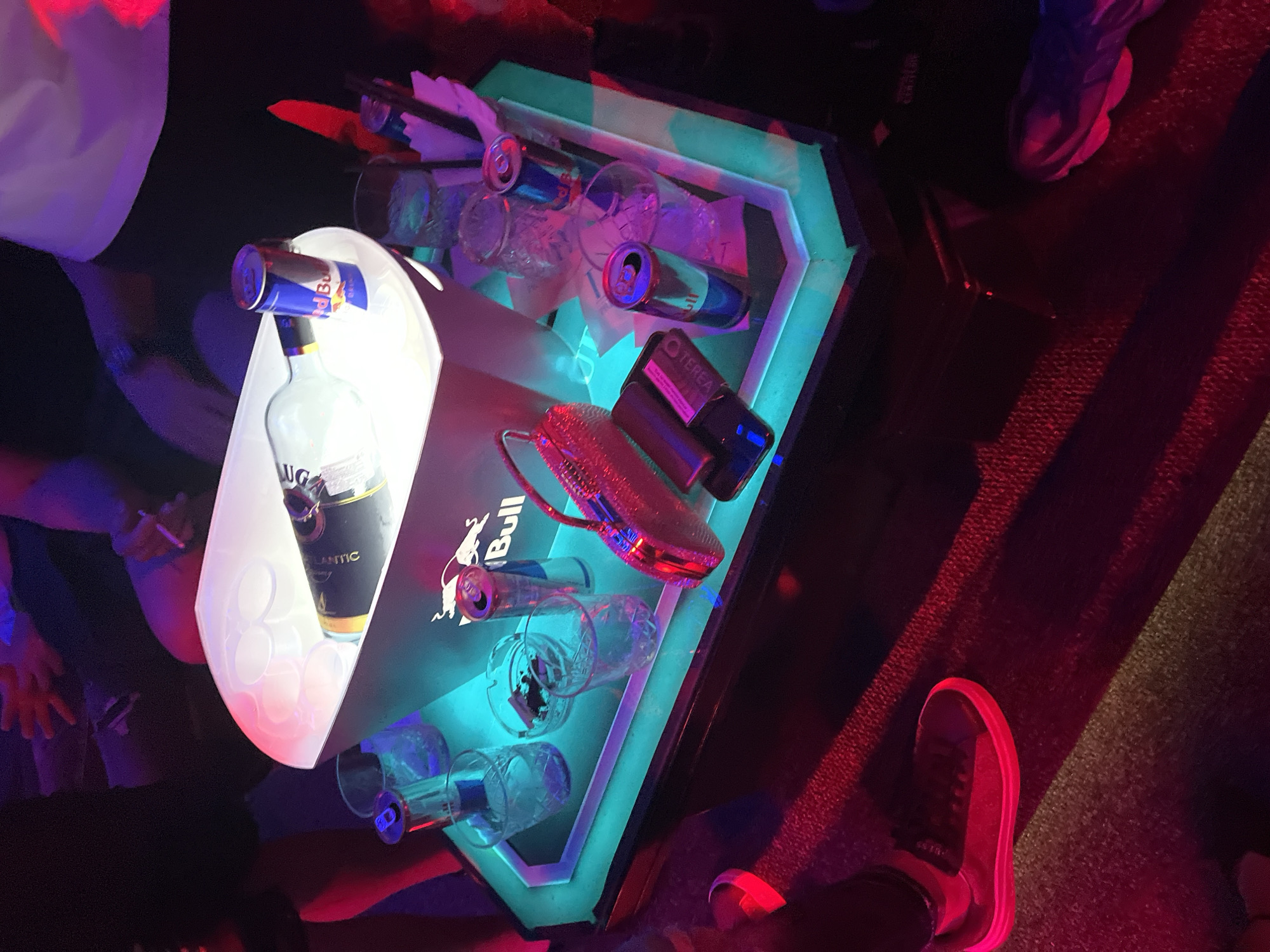 The photograph depicts a nightclub setting, likely in Nesebar, Bulgaria, given the prevalence of Red Bull-branded items, a brand frequently associated with the city. The scene is dimly lit, with strong color contrasts between red, blue, and white light sources. A group of people are gathered around a glowing, multi-level table. The table is adorned with several cans of Red Bull, empty glasses, a bottle of Lug Atlantic liquor, and an accessory resembling a small purse. Other objects include a Tereo-branded item and what seem to be cocktail napkins. Parts of the people standing around the table are visible, including torsos, arms, and legs. Two pairs of sneakers are visible, one being partially visible. The overall impression is that of a busy, active nightclub environment.