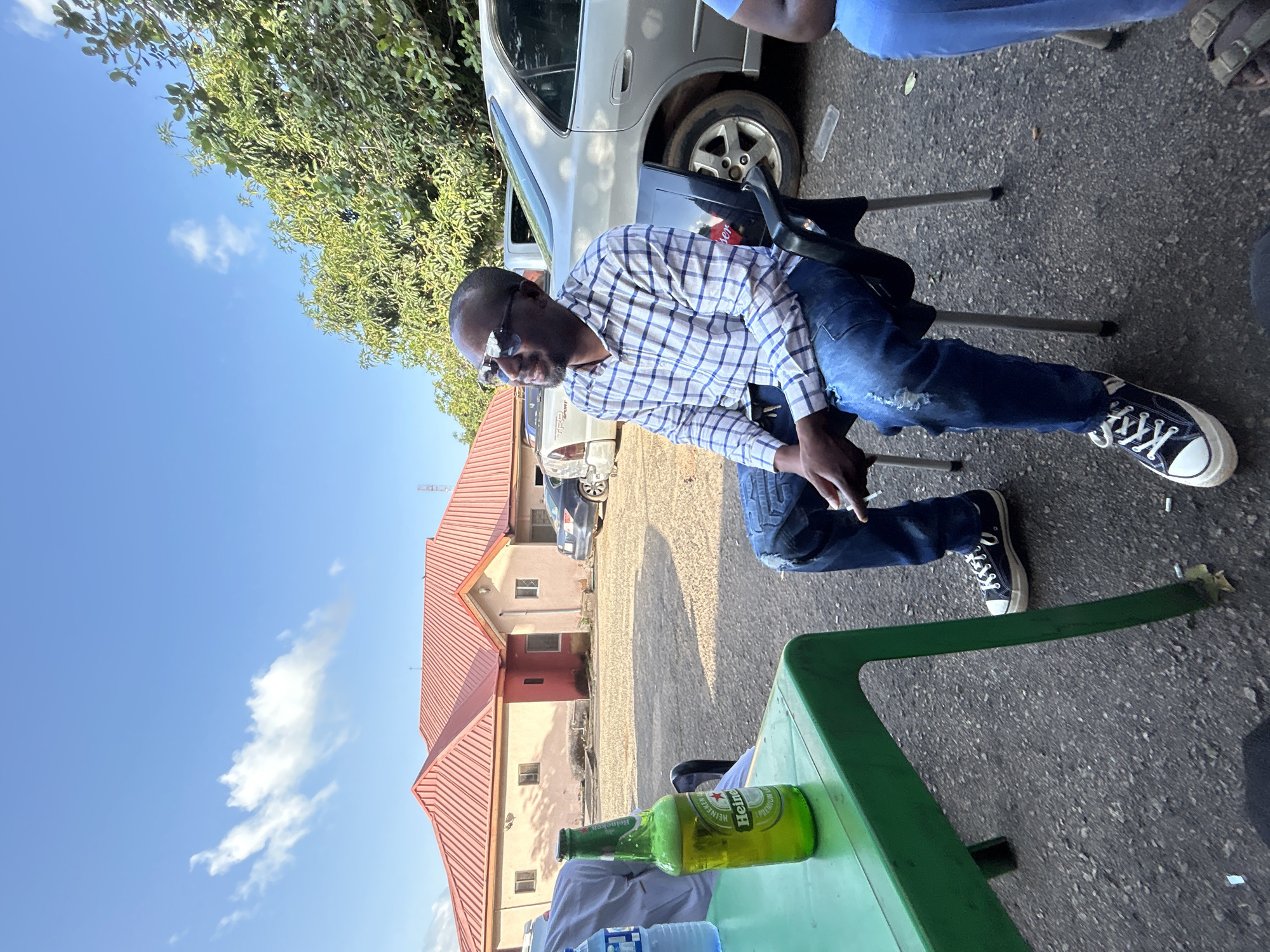 The image depicts an outdoor daytime scene in what is identified as the city of Jos, Nigeria. In the foreground, a dark-skinned male with a short beard and mustache, wearing sunglasses, a blue and white plaid long-sleeve shirt, and ripped blue jeans, is seated on a black plastic chair with metal legs. He is wearing black high-top sneakers with white laces and soles. His left hand holds a small white cylindrical object, possibly a cigarette or vape pen. He is looking upwards and to his right with a slight smile. To his left, a bright green plastic table holds a green Heineken beer bottle, partially full with foam, and a clear plastic water bottle. The lower body of another individual in blue trousers and brown sandals is partially visible to the right of the seated man. A person in a light-colored garment is partially visible behind the green table on the far left. The ground is dark grey, resembling asphalt or gravel, with several white cigarette butts scattered near the table and chairs.
In the midground and background, a silver sedan (resembling a Honda Accord) is parked behind the seated man. Further back, a white pickup truck with "TRD Sport" visible on its tailgate is parked. A single-story building with light brown or beige walls and a prominent red corrugated metal roof stands in the background, featuring several small rectangular windows. Lush green foliage and trees are visible behind the building and vehicles, particularly on the right side of the frame. The sky is bright blue with a few sparse white clouds, and the scene is illuminated by strong daylight, casting clear shadows.