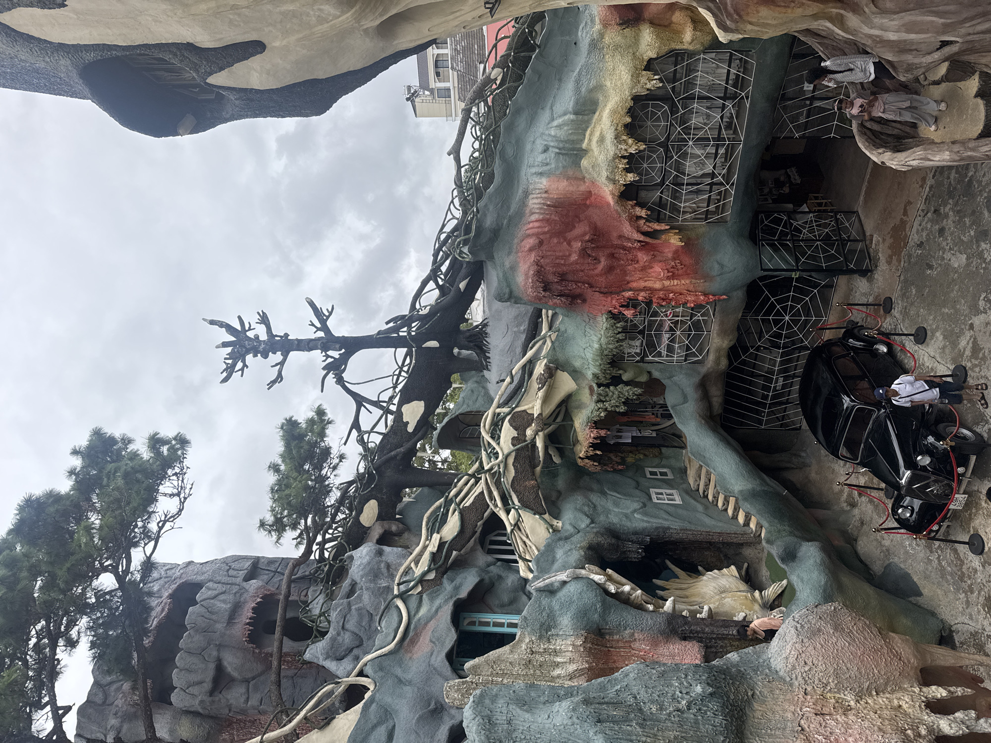 The image displays a section of an eccentrically designed building, characterized by its organic, fantastical architecture, reminiscent of the "Crazy House" in Da Lat, Vietnam. The structure mimics natural forms, appearing as a giant tree, a cave, or an abstract animal, with textured surfaces painted in muted greens, grays, browns, and reddish hues. Artificial roots and branches entwine around the walls, alongside a prominent gnarled tree-like sculpture or real dead tree integrated into the design.
Several people are visible, suggesting a public or tourist attraction. A person in a white top and light pants stands near a vintage-style black car, which is cordoned off with red velvet ropes and gold stanchions, indicating it's an exhibit or photo prop. The car's license plate reads "49A-385.64". Other individuals are partially seen: one in a striped shirt inside an upper opening, another in a pink/peach top near a lower entrance, and a third in light attire within an elevated cave-like space.
Architectural details include white lattice structures resembling spiderwebs covering some window-like openings, and small white-framed windows. Pine trees grow alongside some of the rock-like formations. The sky is uniformly overcast, indicating a cloudy daytime setting. The scene evokes a whimsical and surreal atmosphere, characteristic of the unique location in Da Lat, Vietnam.