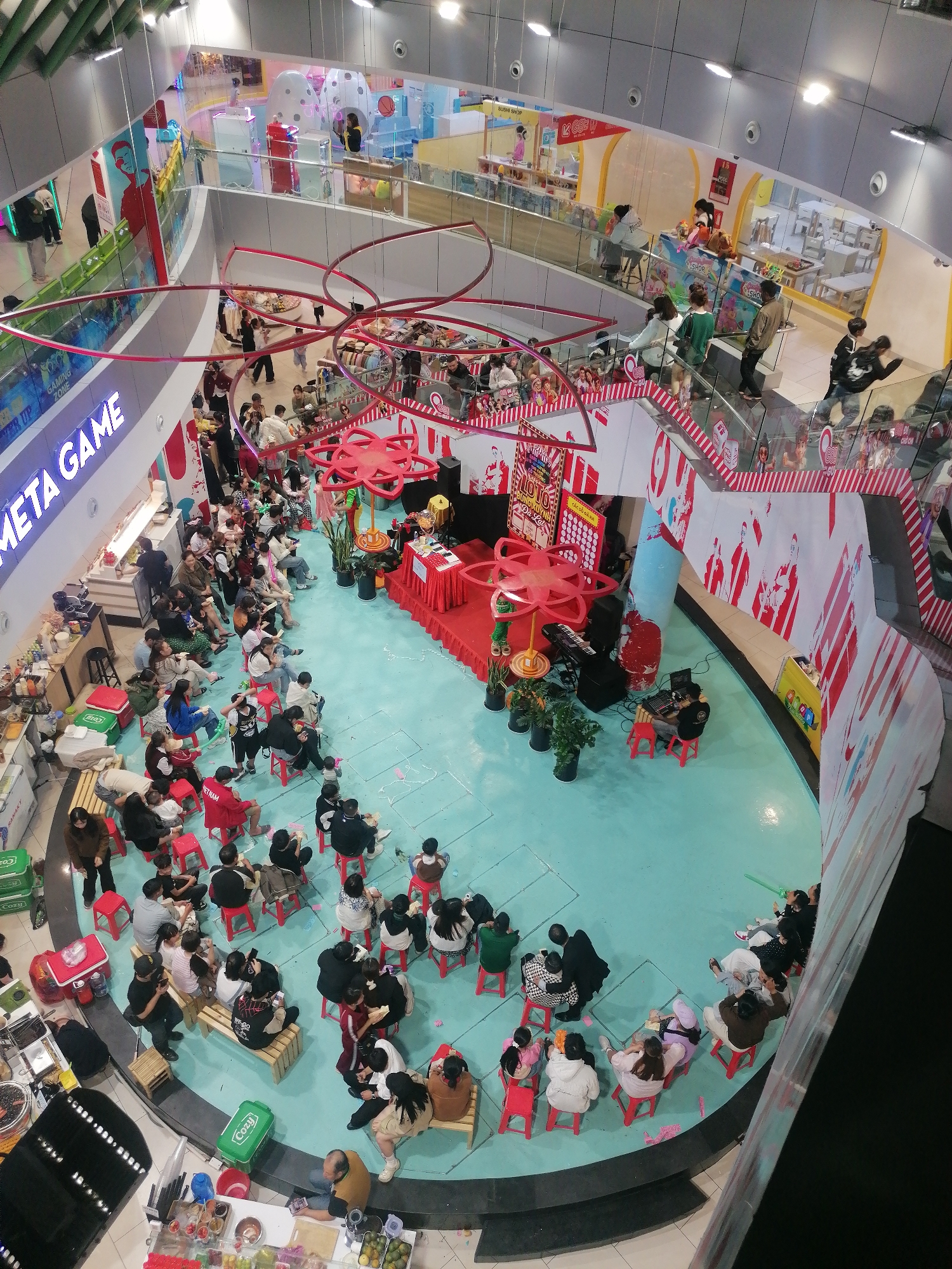 Captured from an elevated perspective, the image shows an event unfolding on a pale blue floor area within a multi-story building, possibly a shopping mall, in Da Lat, Vietnam. A crowd of people, including adults and children, is seated on red stools and benches, facing a stage area where a performance or presentation appears to be taking place. The stage is decorated with red fabrics, flowers, and ornamental features, including what resembles a red flower-shaped structure. Behind the stage are large panels with red and white graphics. Above, suspended circular decorations with red accents hang from the ceiling. Various vendor stands line the perimeter, selling food and drinks, and the image contains signs and logos in English and Vietnamese, including "META GAME," "GAMING ZONE," "SUSHI SHOP," and a brand logo "Cozy." On higher floors, people can be seen looking down at the event from the railing. The environment is illuminated by overhead lighting fixtures.