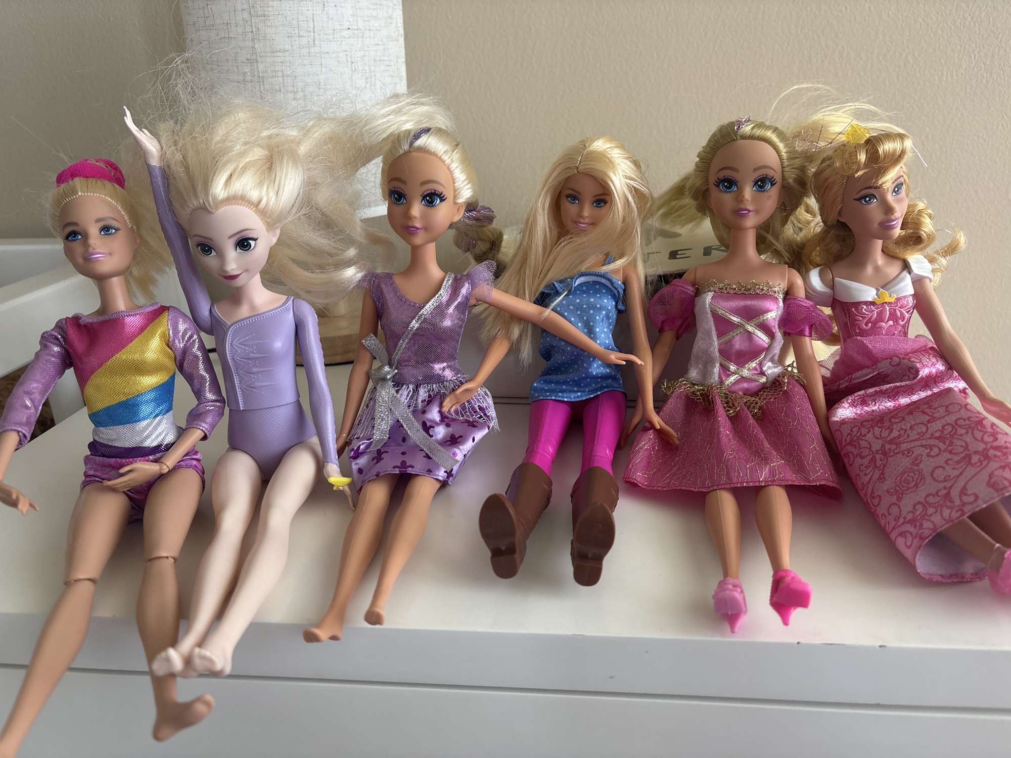 Six dolls are posed sitting on a white shelf. From left to right, the dolls are dressed in various colored outfits: a pink, yellow, and blue leotard with a pink headband; a lavender, sparkly leotard; a purple dress with a sparkly skirt; a blue polka dot top with pink leggings and brown boots; and two pink princess dresses. The dolls have blonde hair, and their expressions are generic to dolls. The background consists of a textured, light-colored wall and a glimpse of a lampshade. The lighting suggests an indoor environment. The image does not contain any elements that would allow for the determination of its location in Dubai, United Arab Emirates.
