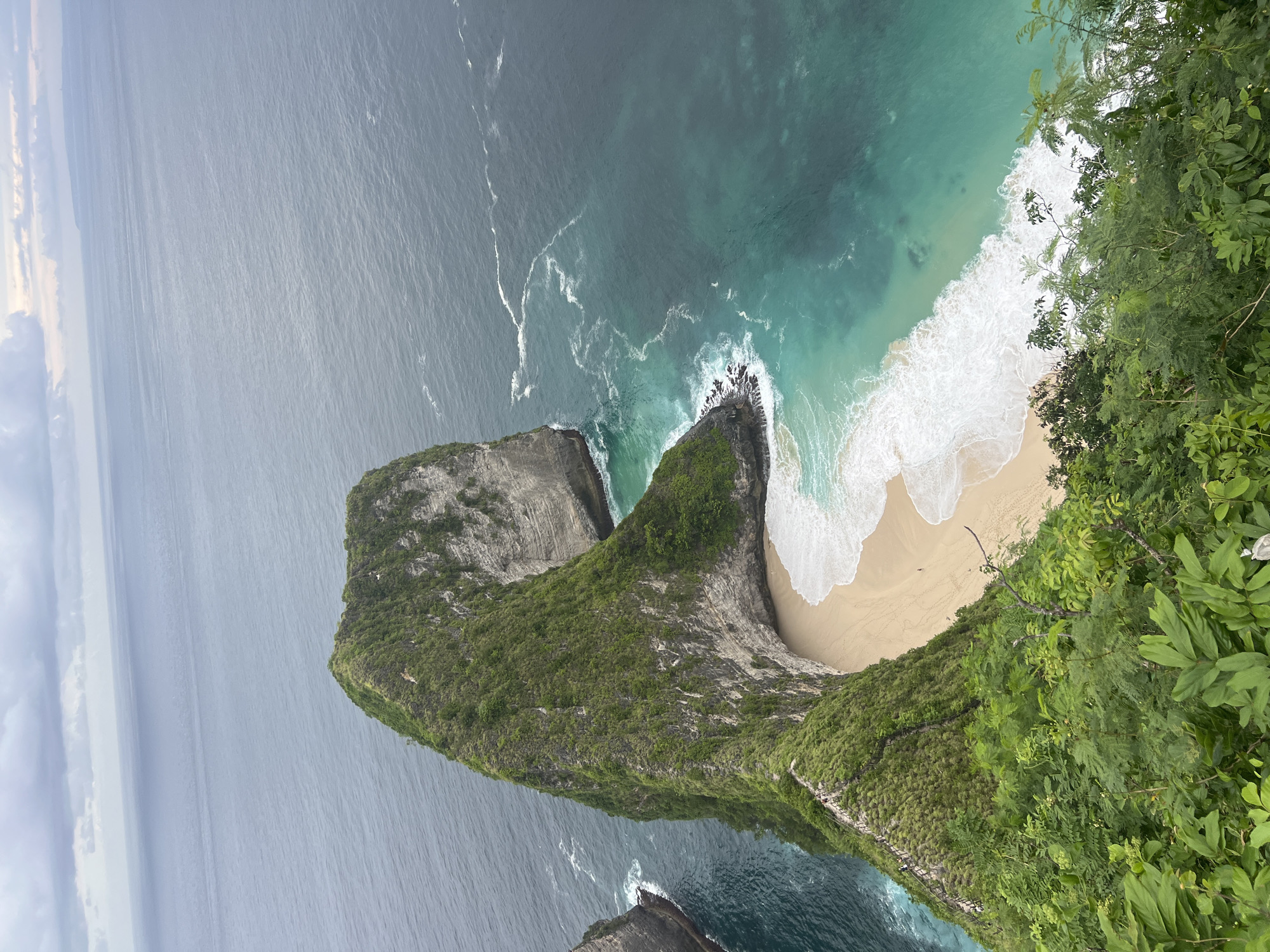 The image shows an aerial view of the Kelingking Beach area in Nusa Penida, Indonesia. The scene features a steep, T-Rex-shaped limestone cliff covered in green vegetation. The cliff overlooks a white-sand beach bordered by turquoise and dark blue water with visible wave activity. The horizon and sky are partially visible in the top left of the image. The scene showcases a clear day at the coast.
