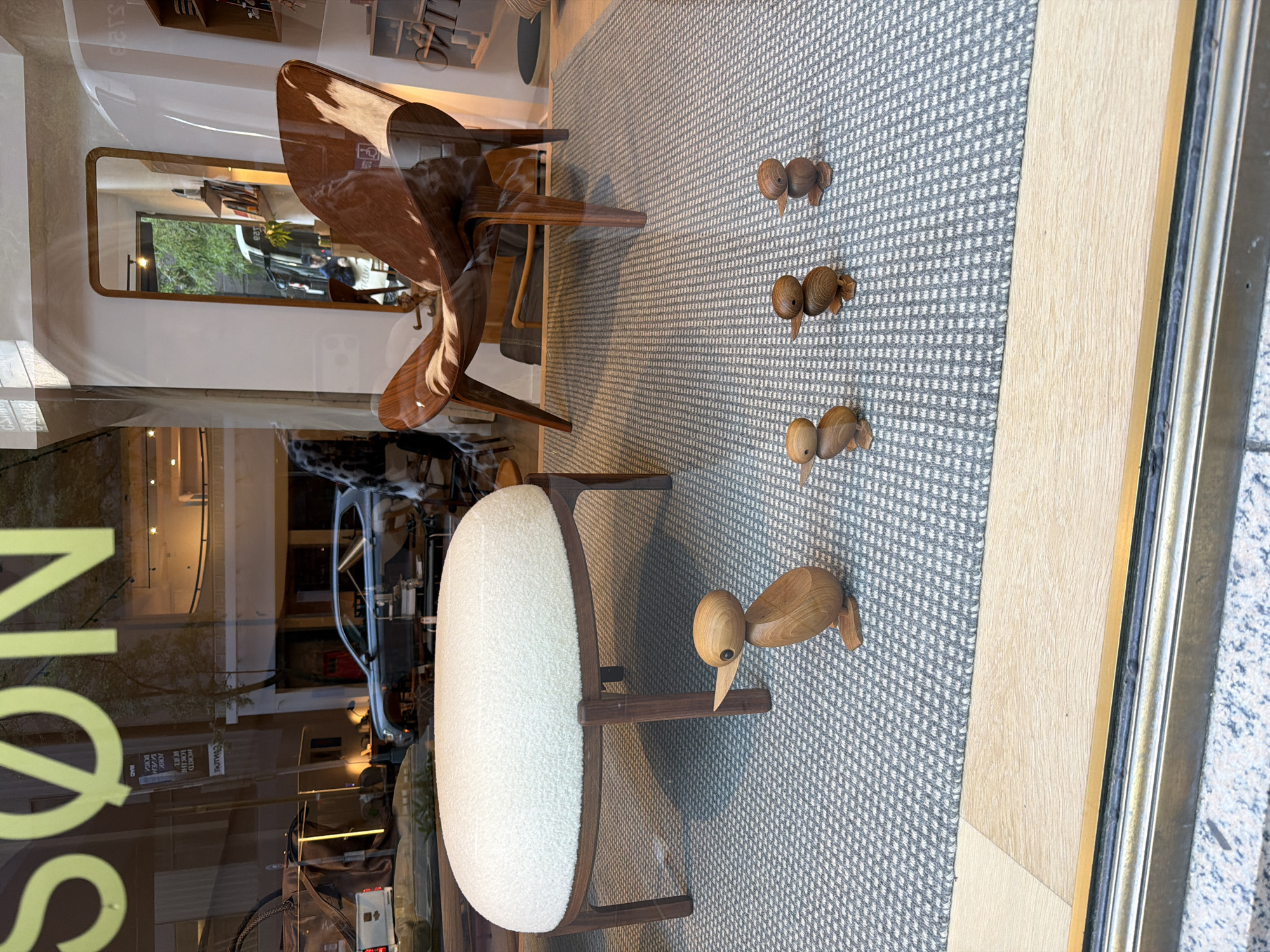 This is a photograph of a store window display on a city street. The window is showing a selection of furniture and decorative items. In the foreground, there is a fluffy white ottoman and a set of four wooden duck figurines arranged in a line. Behind the ottoman, a brown leather chair with a wooden frame is visible. The background of the display shows reflections of the street outside, including cars and buildings, as well as some interior elements of the store such as shelves and lighting. The overall impression is one of a modern and stylish interior design store.