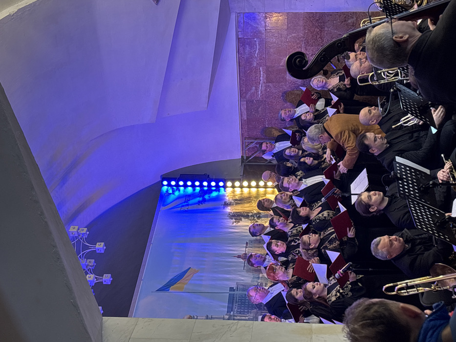 A large choir and orchestra are performing in an underground public space, likely a subway station, in the city of Unknown, Unknown. Approximately 25-30 choir members, dressed in black with gold patterned accents, stand on risers, holding red music binders and singing. In front of the choir, an orchestra of 10-15 musicians, dressed in dark suits, is seated with various instruments including trombones, trumpets, and a double bass, accompanied by black music stands. A male conductor, wearing a brown jacket, stands centrally, directing the ensemble with both hands. The background features a large banner displaying a city skyline and a prominent Ukrainian flag, illuminated by bright blue stage lighting from the left and vertical yellow LED strips on the right. The environment exhibits high, vaulted concrete ceilings, decorative multi-branched lighting fixtures, and a textured reddish-brown tiled wall on the right. The back of a spectator's head is visible in the lower-left foreground.