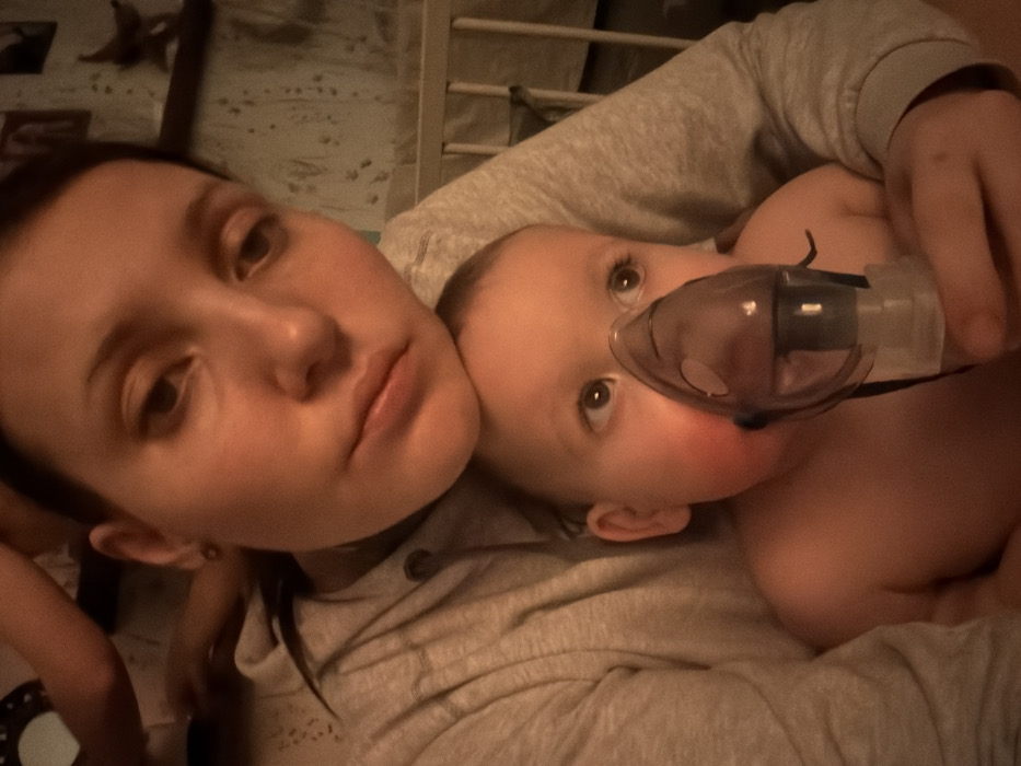A selfie-style shot features a light-skinned woman and a baby lying down. The woman has dark hair, a neutral expression, and is wearing a gray sweatshirt. The baby is shirtless and wearing a nebulizer mask, looking upwards with wide eyes. They appear to be lying on a bed with white sheets and a wooden crib in the background. The overall lighting is dim.