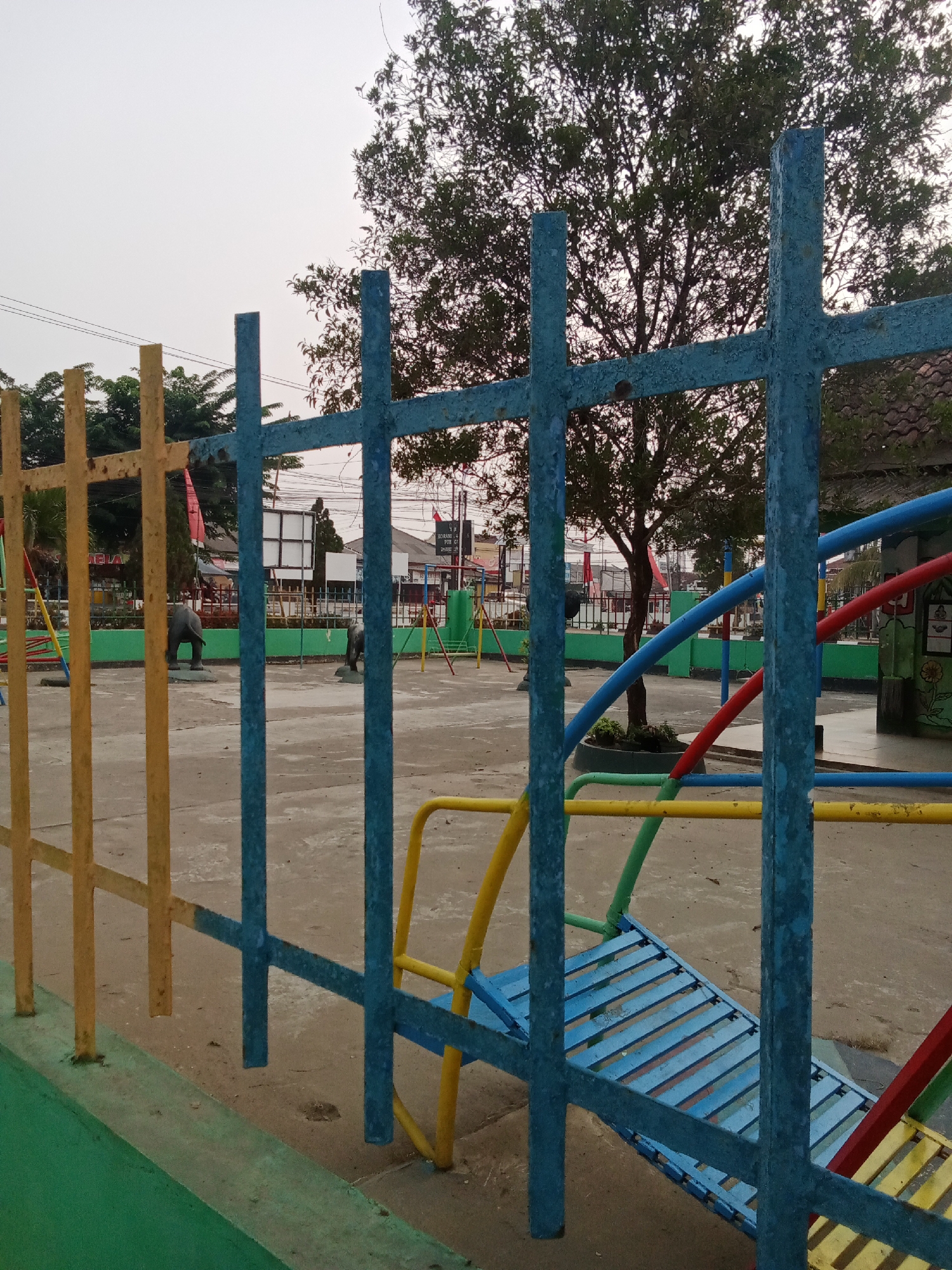 This image depicts an outdoor playground area in Toto Katon, Indonesia, with a view obstructed by a blue metal fence in the foreground. The fence has a criss-cross pattern with vertical and horizontal bars. To the left of the blue fence is a yellow metal fence with vertical bars. Beyond the fences, the playground features a concrete ground surface. In the middle ground, there are two elephant statues, one facing left and the other facing right. There are also various playground equipment structures made of brightly colored metal tubes in red, blue, yellow, and green. A large tree with dense foliage is visible behind the playground equipment. In the background, there are buildings with tiled roofs, and electrical wires crisscross the sky. The overall lighting suggests it is daytime, possibly late afternoon or early morning, with an overcast or hazy sky. There are no people visible in the image.