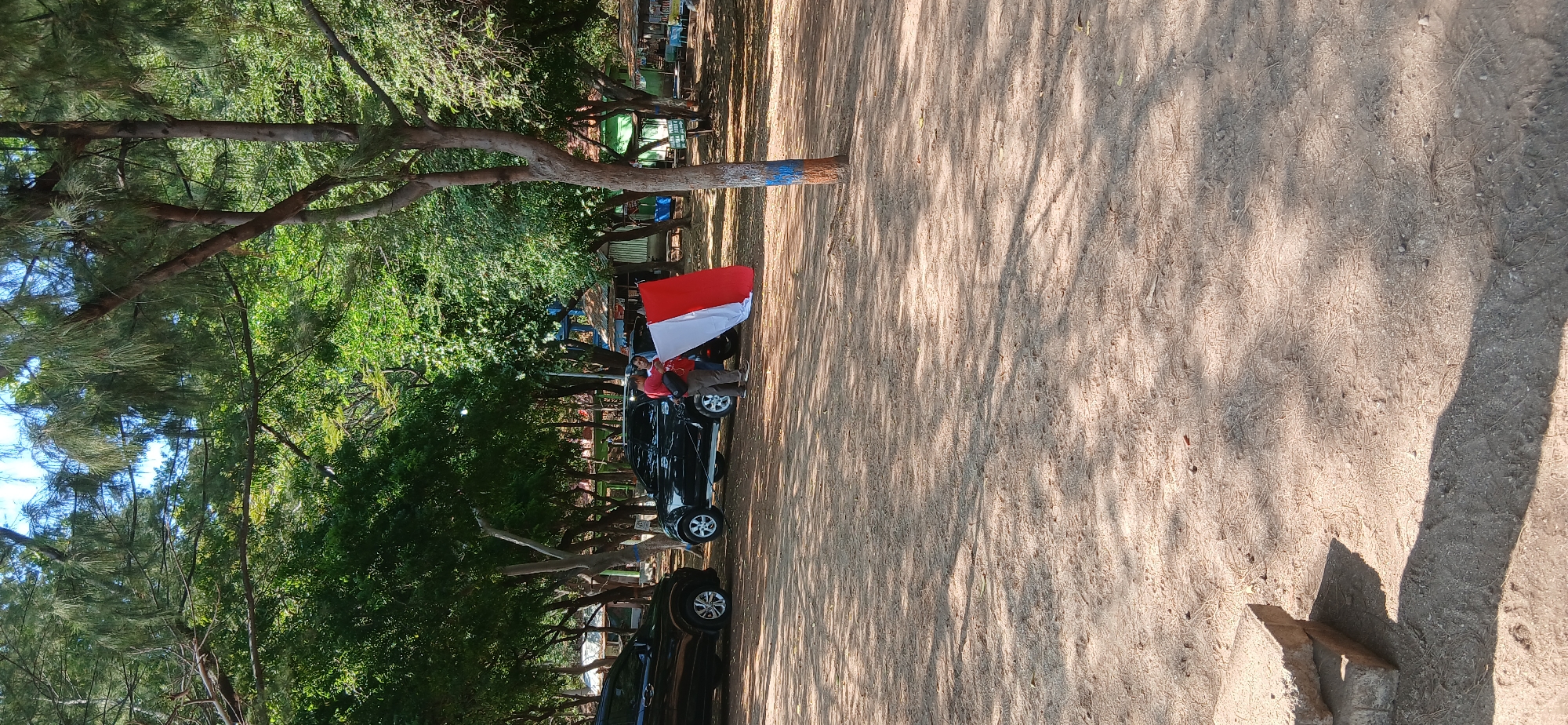 In Banyuglugur, Indonesia, a sandy area is dotted with trees and shadows. Two dark SUVs are parked on the right side of the image. A person wearing a red shirt and a black backpack stands between the two SUVs, holding a red and white flag, which appears to be the Indonesian flag. They are positioned near the rear of the rightmost SUV. In the background, there are several small structures resembling stalls or huts, and more trees. The ground is covered in sand and dry foliage, with shadows cast by the trees. A few pieces of concrete are stacked in the lower left corner of the frame.