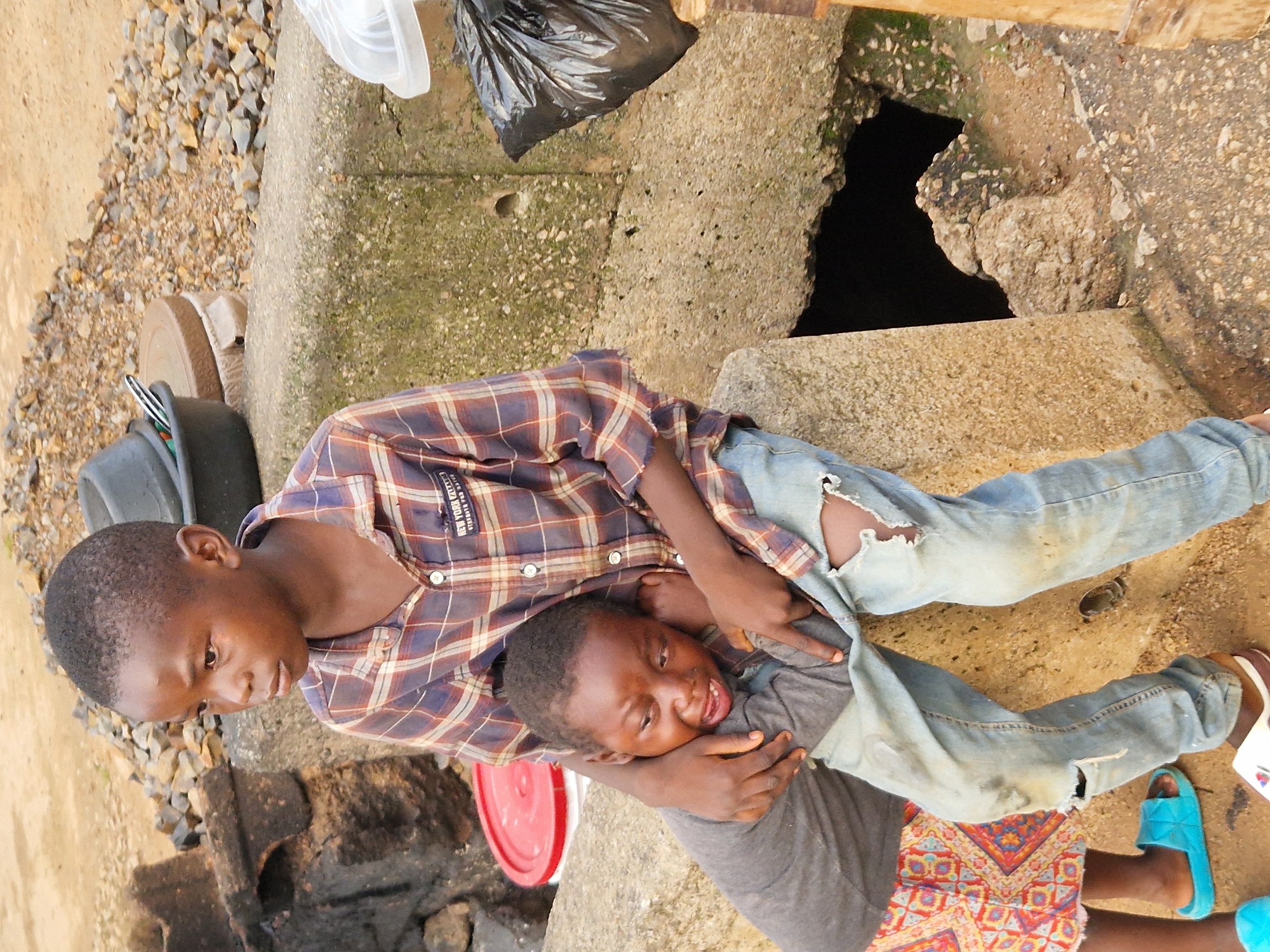 In Kabong, Nigeria, two young children sit on a rough, concrete ledge. The elder child, wearing a plaid shirt and torn jeans, embraces the younger child who is crying and being held tightly. The background shows a dusty, uneven terrain with rocks, debris, and a red bucket. A black plastic bag and a white container are visible to the right of the children. The overall scene depicts a somber moment, hinting at hardship.