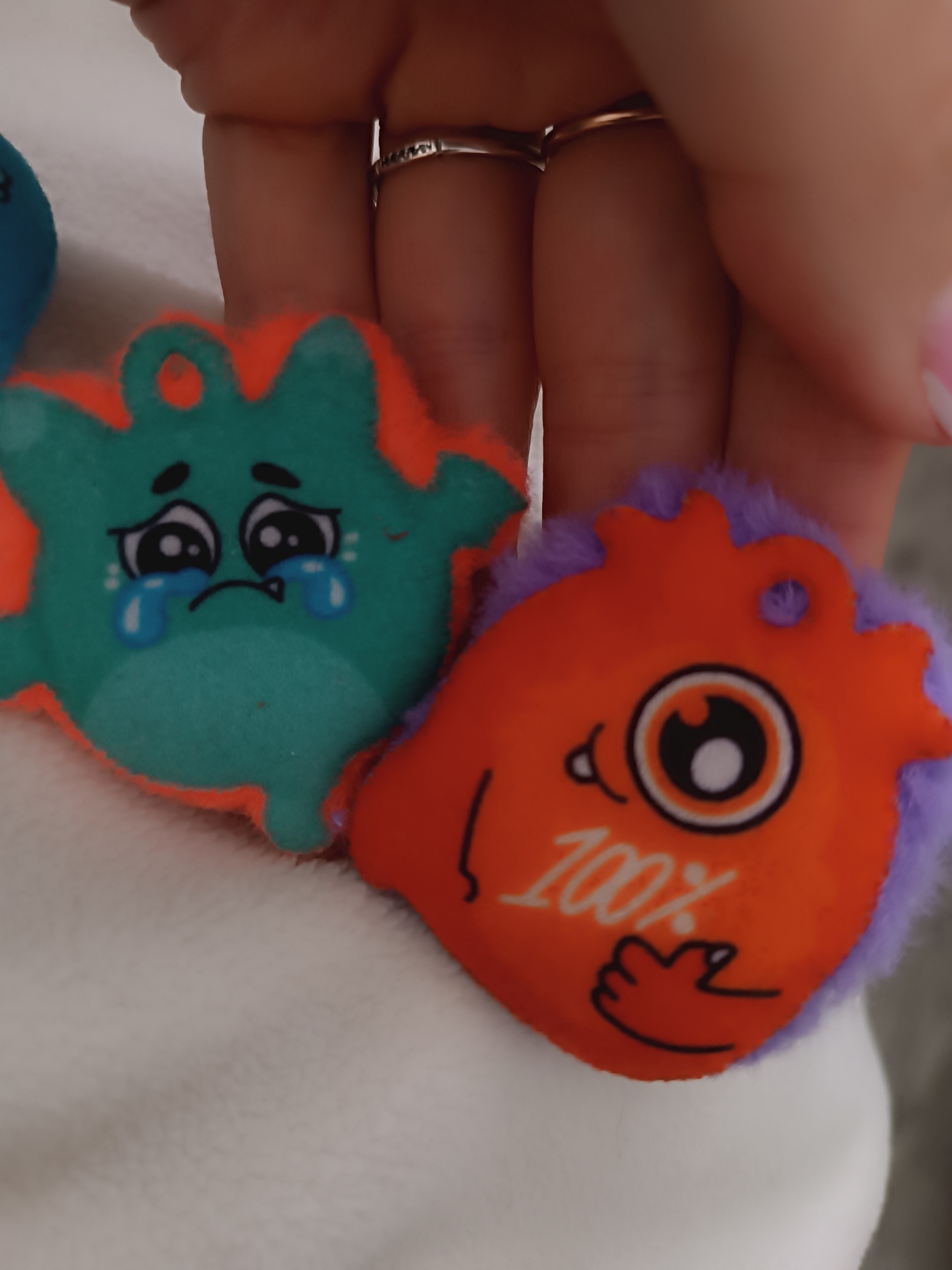The image shows two small, plush keychains being held in a person's hand. The keychain on the left is teal with an orange border and features a cartoon face with large, crying eyes. The keychain on the right is orange with a purple border and features a cartoon face with one large eye and the text "100%" with a hand icon. The person holding the keychains has two rings on their fingers. The background is out of focus and appears to be a white, textured surface. The location is Unknown, Unknown.