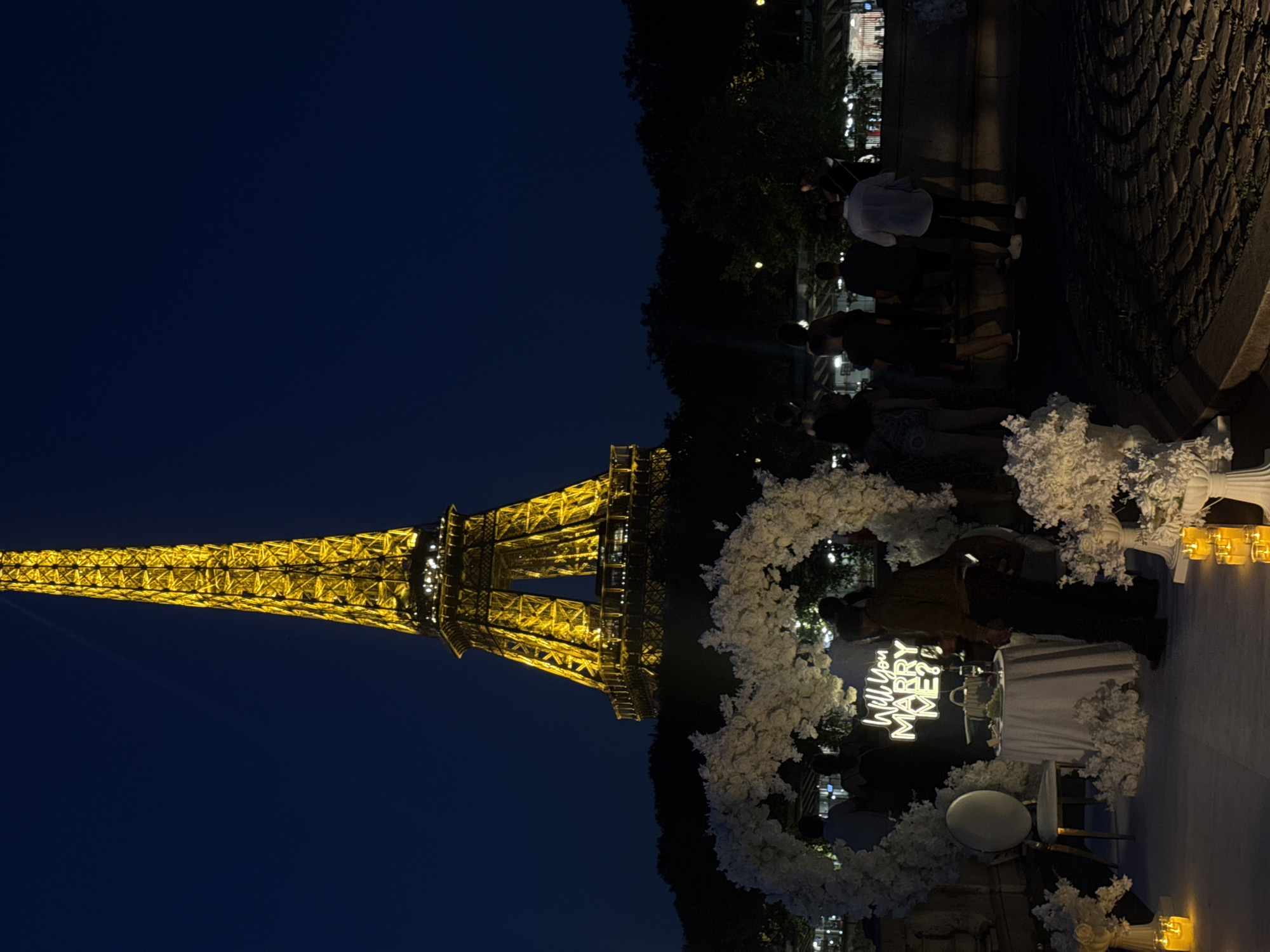 At night in Paris, France, the Eiffel Tower is illuminated and visible in the background. In the foreground, a romantic proposal setup is arranged on a white carpeted platform. A large white floral heart arch frames the scene. Inside the arch, a small round table is draped with a white tablecloth, set with two glasses of champagne and a bowl of fruit. A neon sign behind the table reads "Will You MARRY ME?". To the left of the table, a single white chair with a gold frame is placed. Several people are standing in front of the setup, observing or participating in the event. More floral decorations and lit candles are placed around the platform. The ground in the foreground is paved with cobblestones.