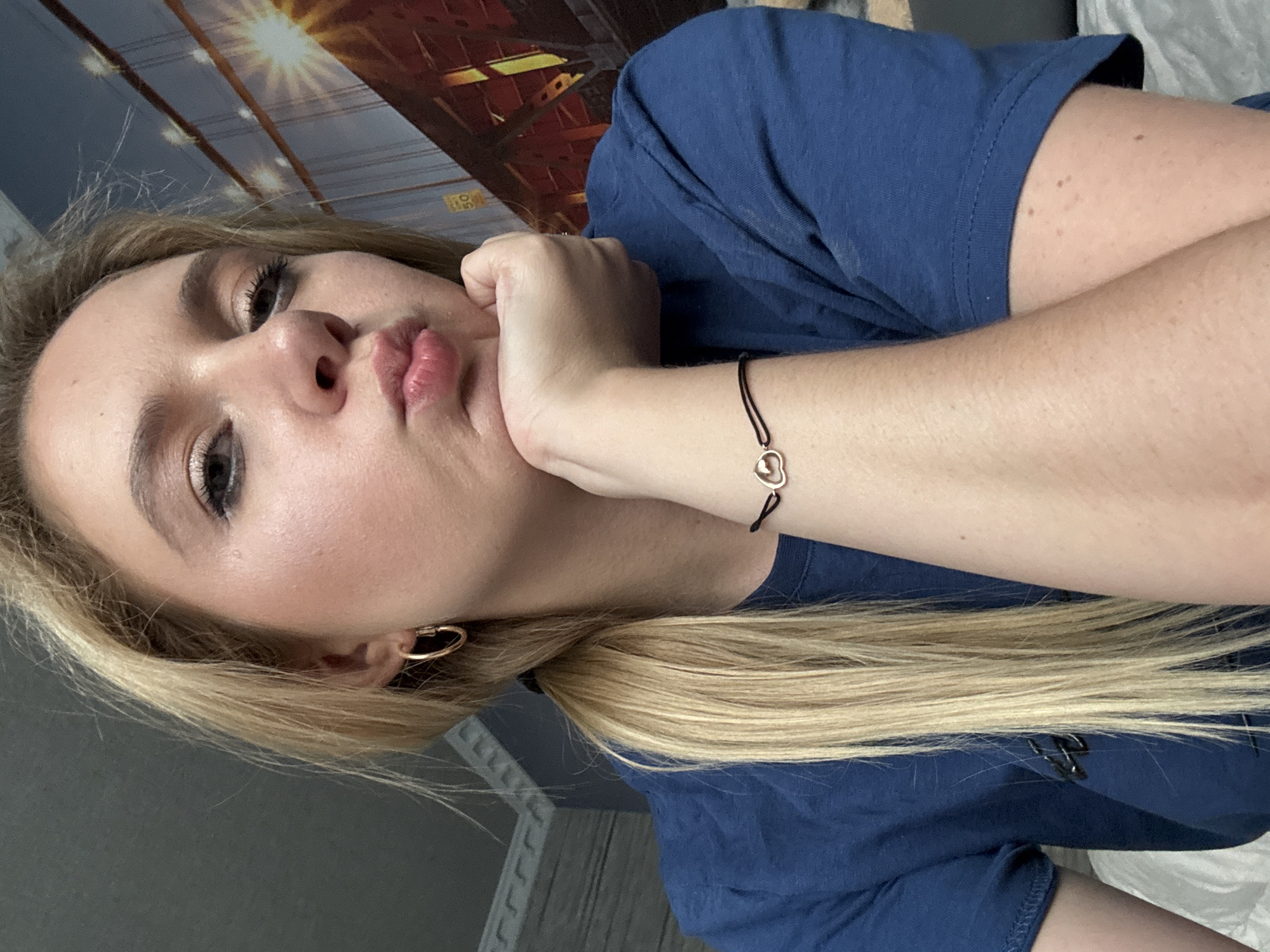 A young Caucasian female with blonde hair styled in a ponytail and wearing a blue t-shirt is shown from the shoulders up. She is looking at the camera with her chin resting on her hand, her lips pursed. She is wearing gold hoop earrings and a black string bracelet with a gold heart charm on her wrist. The background features a wall with ornate trim and a large image of a suspension bridge with a sunset or sunrise behind it. The scene appears to be indoors, possibly a bedroom or living area.