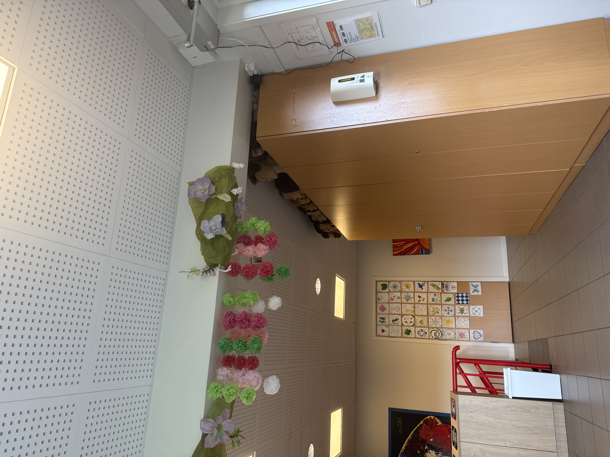 The image depicts an interior scene, likely a classroom or common area, featuring a white perforated ceiling with recessed lighting. Decorative paper pom-poms in various shades of pink, green, and white are suspended from a green fabric garland draped across the top of a light brown wooden cabinet. The cabinet is tall and appears to have multiple doors, with a white soap dispenser mounted on the right-hand side.

Peeking over the top of the cabinet, several mannequin heads are visible, arranged in a row. To the left of the cabinet, a section of a lighter colored wall is visible, adorned with a grid of small, square tiles, each featuring a unique drawing or pattern. Adjacent to this tiled wall is a red railing leading to what appears to be a stairwell, with a white trash bin positioned below it. A light wooden counter is also visible to the far left.

The floor is tiled with square, grey tiles. The overall impression is one of a space that has been decorated for a festive occasion or for artistic display. No specific landmarks or details in the image definitively place it in Skatval, Norway, though it could be a room within a school, community center, or other public building in that location.