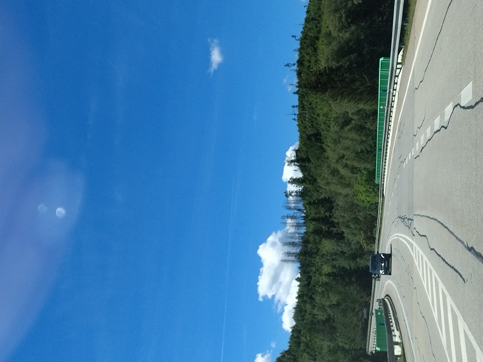 The image depicts a highway curving through a forested mountainous landscape, likely in the vicinity of Strážný, Czechia, given the provided context. The sky is a bright, clear blue with a few wispy clouds and two bright circular artifacts, possibly lens flare or reflections. A dense green forest covers the hillsides, with some areas showing patches of dead, bare trees, suggesting past deforestation or natural events. The highway itself is paved with visible cracks and markings. On the road, a dark-colored SUV is driving away from the viewer, and a motorcycle is visible behind it. Guardrails line the sides of the highway, with a distinctive green barrier visible on the right side of the road. The overall scene suggests a sunny day on a mountain road.