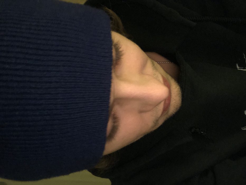 A close-up shot shows a person's face mostly sideways, wearing a dark blue knit hat pulled low over the forehead. Visible facial features include an eye, nose, mouth, and some facial hair. The person wears a black hoodie with a glimpse of a silver beaded necklace visible at the neck. The image appears to have low lighting or was taken with a low-quality camera.