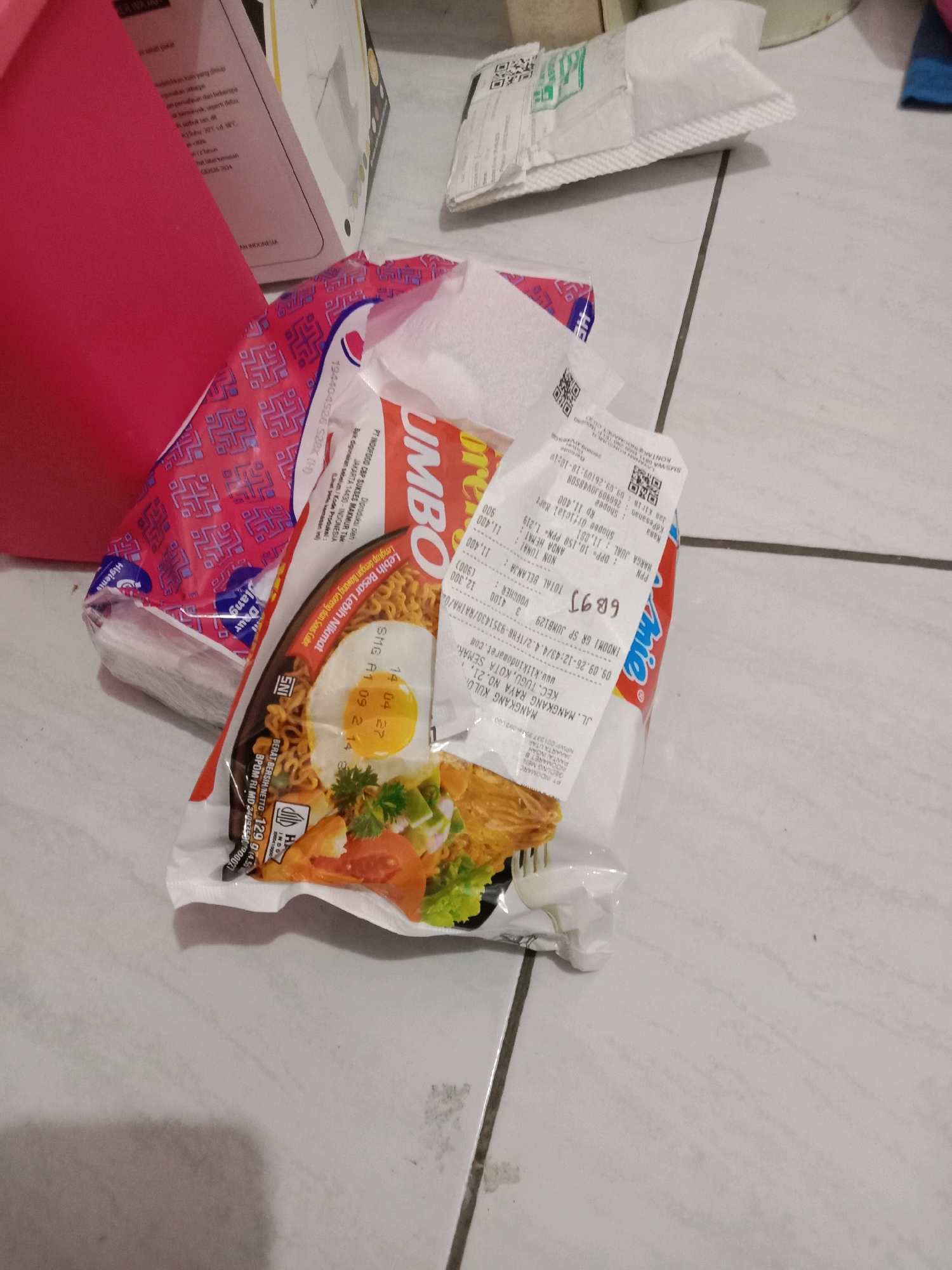 The image shows a close-up, eye-level shot of instant noodles and a receipt on a tiled floor. The noodles are in a red and white package with an image of a fried egg and vegetables. A white receipt with black text is partially visible, indicating a purchase from "PT. INDOFRESH" in "JL. MANGKANG KAYA NO. 21, KEC. TUGU KOTA SEMARANG". The floor is light-colored tile with faint gray grout lines. The scene is indoors.
