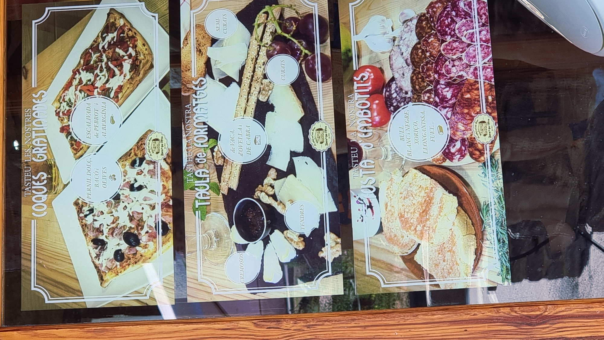 The image is a photograph of a menu display board for a restaurant in Rupit, Spain. The display features three sections showcasing different food items.
The top section, titled "TASTEU LES NOSTRES COQUES GRATINADES," displays two types of gratinated flatbreads. The one on the left is topped with ham, bacon, and olives. The one on the right appears to be an escalivada, with roasted red peppers and eggplant.
The middle section, titled "TAULA de FORMATGES," shows a cheese platter. Various types of cheese are arranged on a dark surface, accompanied by crackers, walnuts, grapes, and a small bowl of jam. The cheeses are labeled as "CREMOSOS" (creamy), "TENDRES" (tender), "DE VACA, D'OVELLA i DE CABRA" (from cow, sheep, and goat), "SEMI-CURATS" (semi-cured), and "CURATS" (cured).
The bottom section, titled "TASTA D'EMBOTITS," presents a selection of cured meats. The display includes bread, tomatoes, garlic, and an assortment of sliced sausages. The sausages are described as "BULL BLANC i NEGRE, XORIÇO, LLONGANISSA, FUET..." (white and black sausage, chorizo, longanissa, fuet).
Each section includes a small circular emblem with text, likely indicating the restaurant's name and location. The overall presentation suggests a rustic and traditional culinary offering.
