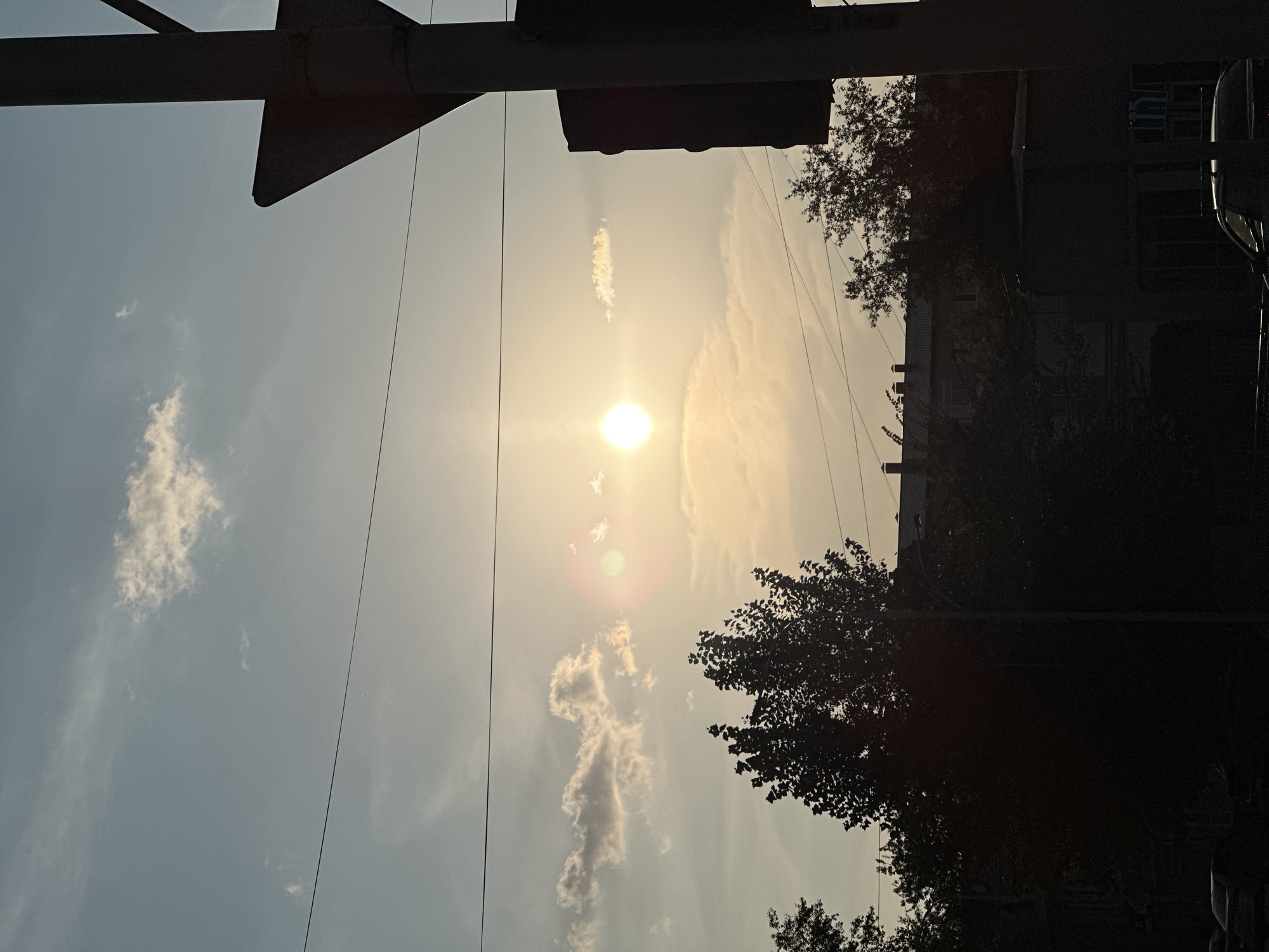 This image depicts a view of the sky with the sun shining brightly, creating a lens flare. The sun is partially obscured by thin, wispy clouds. In the foreground, silhouetted against the bright sky, are the dark shapes of trees and buildings, suggesting an urban or semi-urban setting. A traffic light or similar overhead structure is visible in the top left corner. The image is taken from a low angle, looking upwards. The lighting suggests it is either early morning or late afternoon, with the sun low on the horizon. The presence of clouds indicates a partly cloudy sky, and the overall atmosphere is serene. No people are visible in the frame. The crops provided offer closer views of the overhead structure, the sun and clouds, and the silhouetted trees and buildings. There is no visible text in the image.