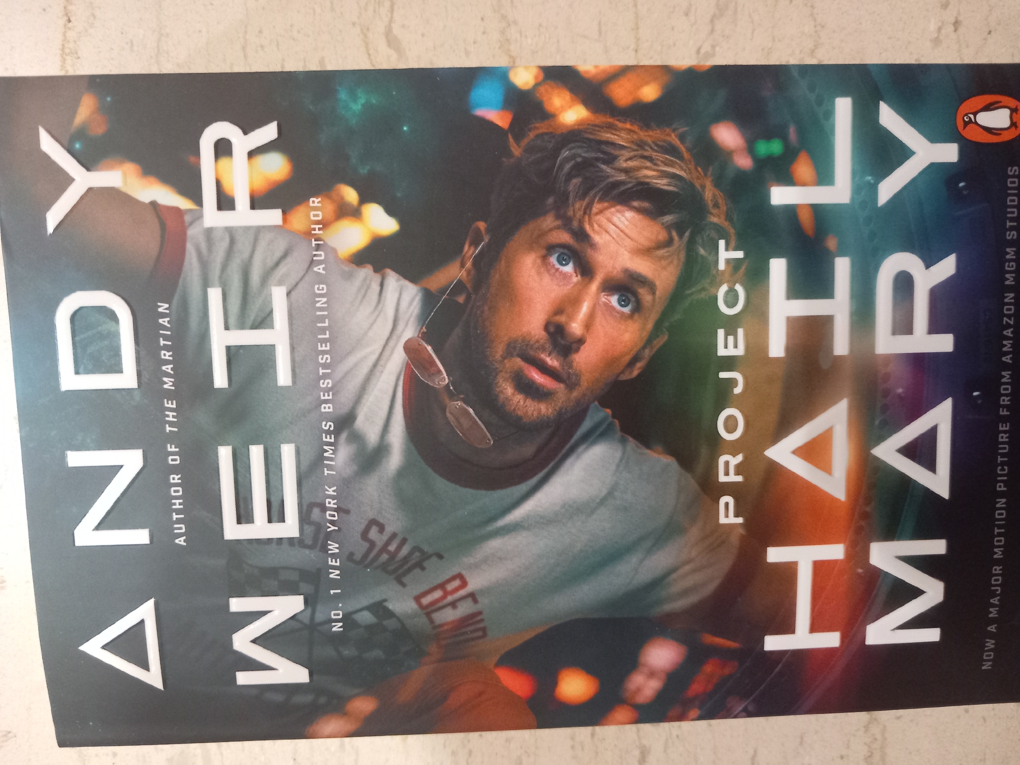 The image is a close-up, slightly angled shot of the cover of the book "Project Hail Mary" by Andy Weir. The book cover features a man's face tilted to the side, with his blue eyes looking directly at the camera. He has short brown hair and is wearing sunglasses perched on his head. He is wearing a light-colored t-shirt with the words "SHOE BEND" printed on it in a stylized font. The title "PROJECT HAIL MARY" is prominently displayed in large white letters across the bottom half of the cover, with "PROJECT" in a smaller font above the larger letters. "ANDY WEIR" is written in large white letters at the top, and below it, "AUTHOR OF THE MARTIAN" and "NO. 1 NEW YORK TIMES BESTSELLING AUTHOR." The background is dark with streaks of colorful light, suggesting a cosmic or futuristic setting. The bottom edge of the cover shows the text "NOW A MAJOR MOTION PICTURE FROM AMAZON MGM STUDIOS" and a small orange penguin logo, likely indicating the publisher. The book is lying flat on a textured, light-colored surface, possibly a countertop or floor.