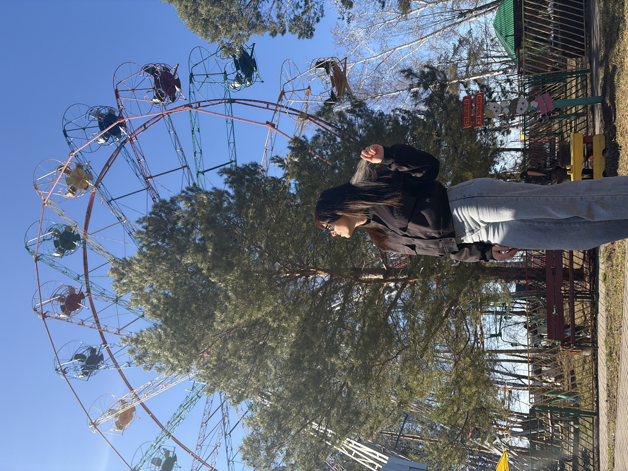 This brightly lit, outdoor image captures a scene at what appears to be an amusement park or recreational area in Chaikovsky, Russia.
The central subject is a young woman, seen from the waist up in profile, positioned on the right side of the frame. She has long, dark hair, wears a black jacket or top, and light-colored jeans, and is looking slightly upwards and to the right.
Dominating the left and central parts of the image is a large Ferris wheel featuring a multi-colored structure with gondolas or frames in shades of red, green, blue, and yellow. The sky is a clear, vibrant blue, indicating sunny weather during daytime, likely morning or early afternoon.
Various trees are interspersed throughout the park, including tall evergreen pines with dense foliage and some bare deciduous trees, suggesting late winter or early spring. In the background, to the right of the woman, other amusement park structures or playground equipment are visible, alongside a fence and a small building with a green roof. A paved pathway or sidewalk runs along the far right foreground.
Notable visible text includes two red signs in the background with white or light-colored Cyrillic letters. The clearer sign prominently reads "ЦИРК!" (Circus!). Below these signs are cut-out figures resembling animated characters, specifically Masha and the Bear. The overall impression is one of a calm and pleasant day at a park.