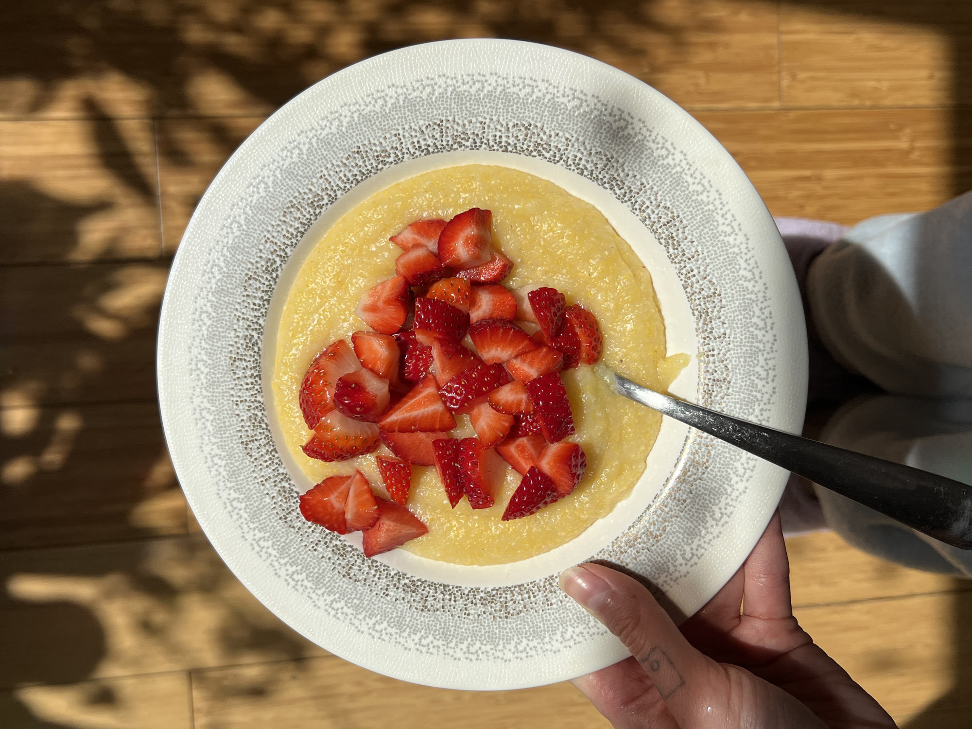A person holds a plate of yellow polenta topped with chopped strawberries, garnished with a silver spoon on a hardwood floor under sunlight filtering through branches, presumably indoors. The plate has a white and silver speckled pattern. The hand holding the plate has pale skin and a small tattoo. The scene does not contain enough information to locate the specific location of the city of Palilula Urban Municipality, Serbia.